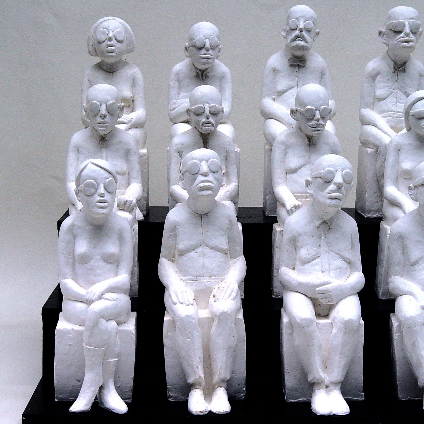 این تصویر یک گروه از مجسمه‌های سفید رنگ را نشان می‌دهد که به شکل انسان‌ها نشسته‌اند. آنها در دو ردیف و به تعداد ده نفر قرار دارند. هر مجسمه ویژگی‌های خاصی دارد، مانند عینک و حالت‌های متفاوت صورت. این مجسمه‌ها ظاهری ساده و انتزاعی دارند و در مجموع حس سکوت و تأمل را منتقل می‌کنند. فضای این اثر هنری می‌تواند به تفکر در مورد زندگی و ارتباطات انسانی اشاره داشته باشد.