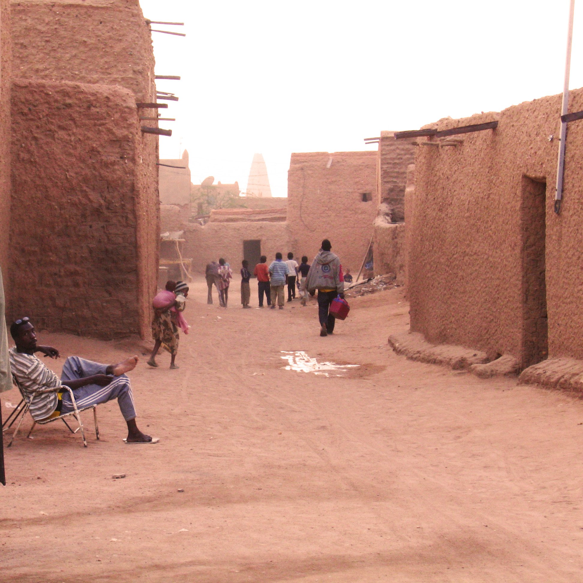 L'image montre une rue dans un village traditionnel, probablement situé dans une région désertique. On y voit des maisons en terre, avec des murs de couleur ocre. Plusieurs personnes déambulent tranquillement dans la rue, certaines s'arrêtent pour discuter. À gauche, un homme est assis sur une moto, tandis qu'un autre se repose sur une chaise. L'atmosphère est calme et chaleureuse, avec un éclairage doux peut-être dû au coucher du soleil. Les ombres et la poussière soulignent le caractère rustique du lieu.