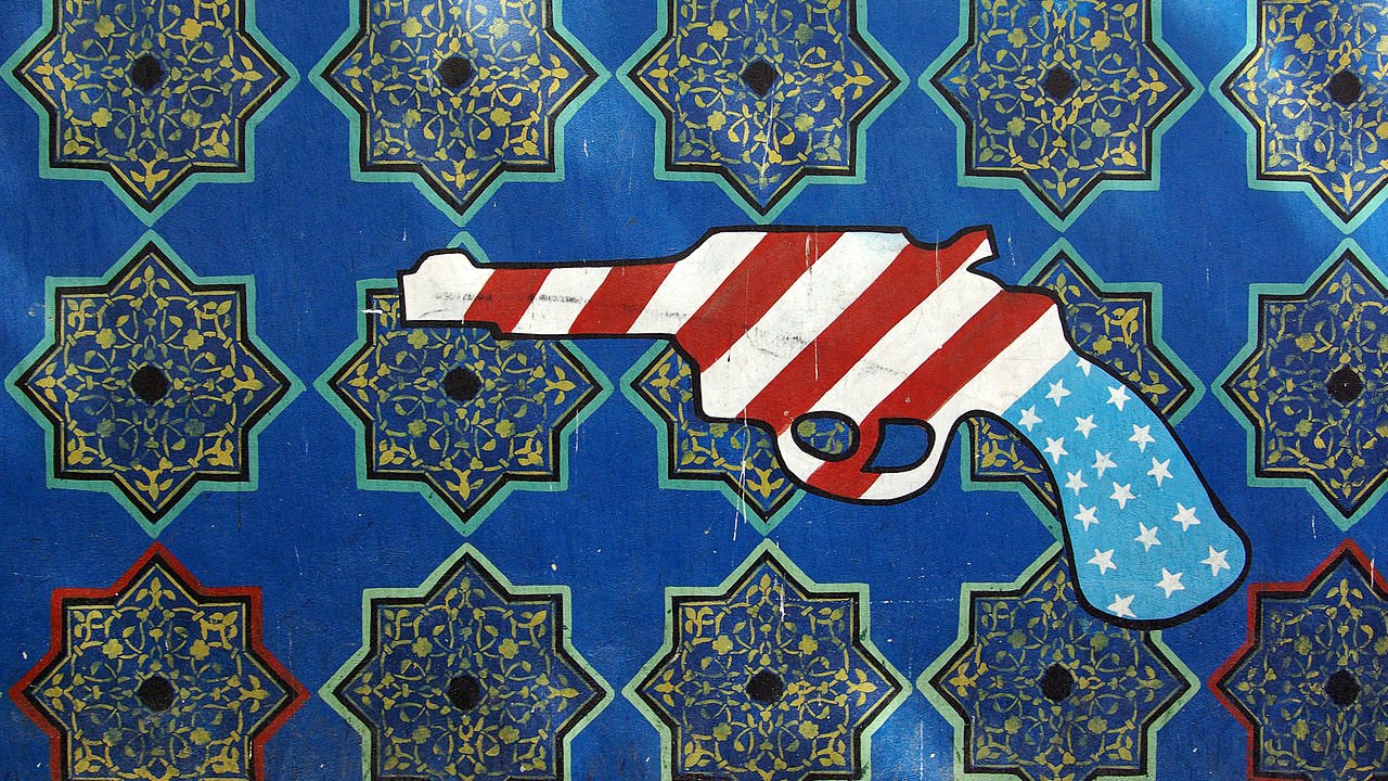 L'image représente un pistolet stylisé recouvert des motifs du drapeau américain, avec des rayures rouges et blanches et des étoiles blanches sur un fond bleu. Le pistolet est placé sur un arrière-plan orné de motifs géométriques complexes aux couleurs vives, qui évoquent des designs traditionnels. Le contraste entre le pistolet et l'arrière-plan artistique souligne un message potentiellement provocateur.