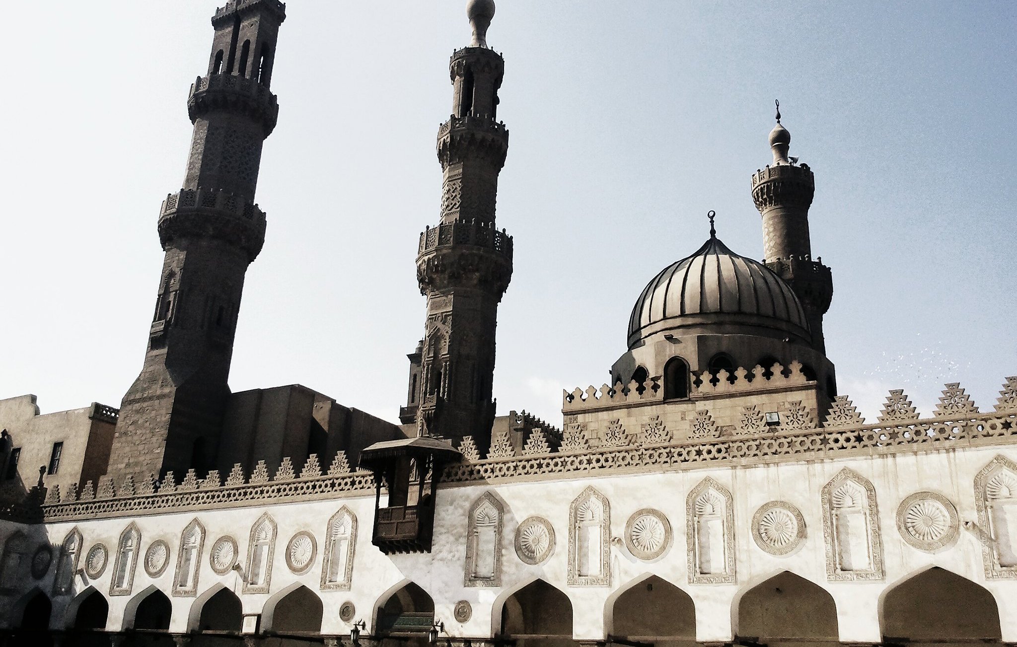 تظهر في الصورة مجموعة من المعالم المعمارية الإسلامية، حيث يتواجد مسجد كبير ذو قبتين ومئذنتين بارزتين. يبرز تصميم المسجد بتفاصيله المعمارية الفريدة، مثل النقوش والزخارف على الجدران. السماء خلف المبنى صافية، ما يضفي جواً من الهدوء على المشهد. تعكس الصورة الطابع التاريخي والثقافي للعمارة الإسلامية.