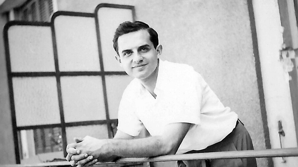 در این تصویر، مردی جوان به نظر می‌رسد که در حال لبخند زدن است. او با آستین‌های کوتاه و پیراهن سفیدی لباس پوشیده و در بالکن یا جایی مشابه ایستاده است. دستانش را بر روی نرده‌های بالکن گذاشته و به دوربین نگاه می‌کند. پس‌زمینه تصویر شامل دیواری ساده و پنجره‌ای با شیشه‌های مشبک است. جو تصویر دوستانه و صمیمانه به نظر می‌رسد.