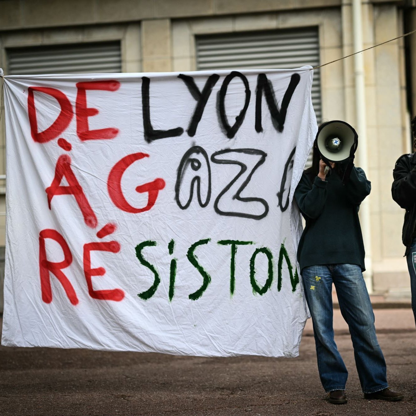 L'immagine mostra due persone che reggono un grande striscione bianco con le parole "DE LYON À GAZA RÉSISTON" scritte in caratteri grandi e colorati. Le due persone hanno in mano dei megafoni e sembrano partecipare a una manifestazione o a un evento di protesta. Lo sfondo suggerisce un contesto urbano, probabilmente in una zona pubblica.