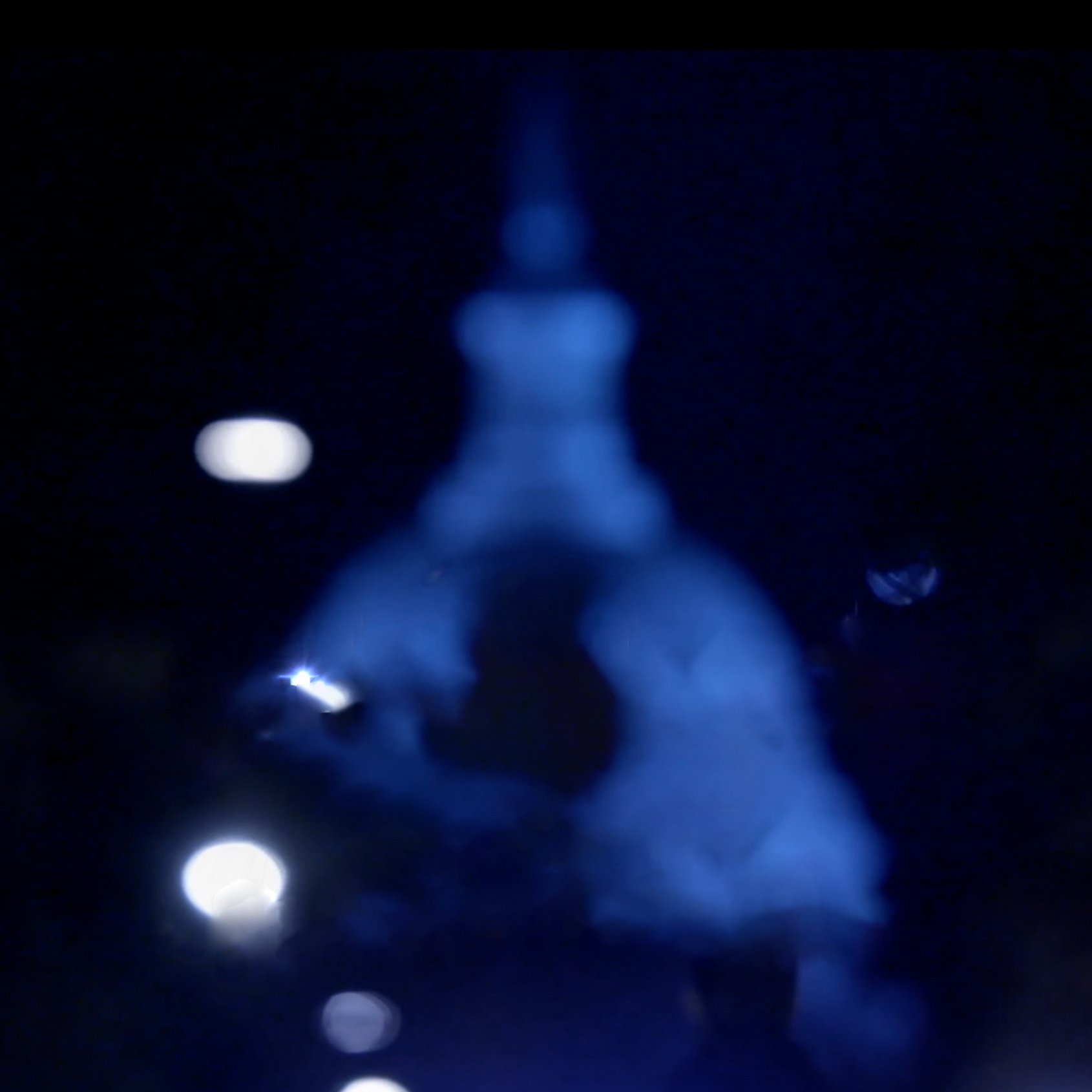 L'image montre une scène nocturne avec un éclairage diffus. Au centre, on aperçoit une silhouette floue, probablement d'un bâtiment ou d'une structure, avec un dôme et une pointe. Les lumières autour ajoutent une ambiance mystérieuse et éthérée, créant un contraste entre l'obscurité environnante et les sources lumineuses. L'atmosphère semble calme et légèrement mélancolique.