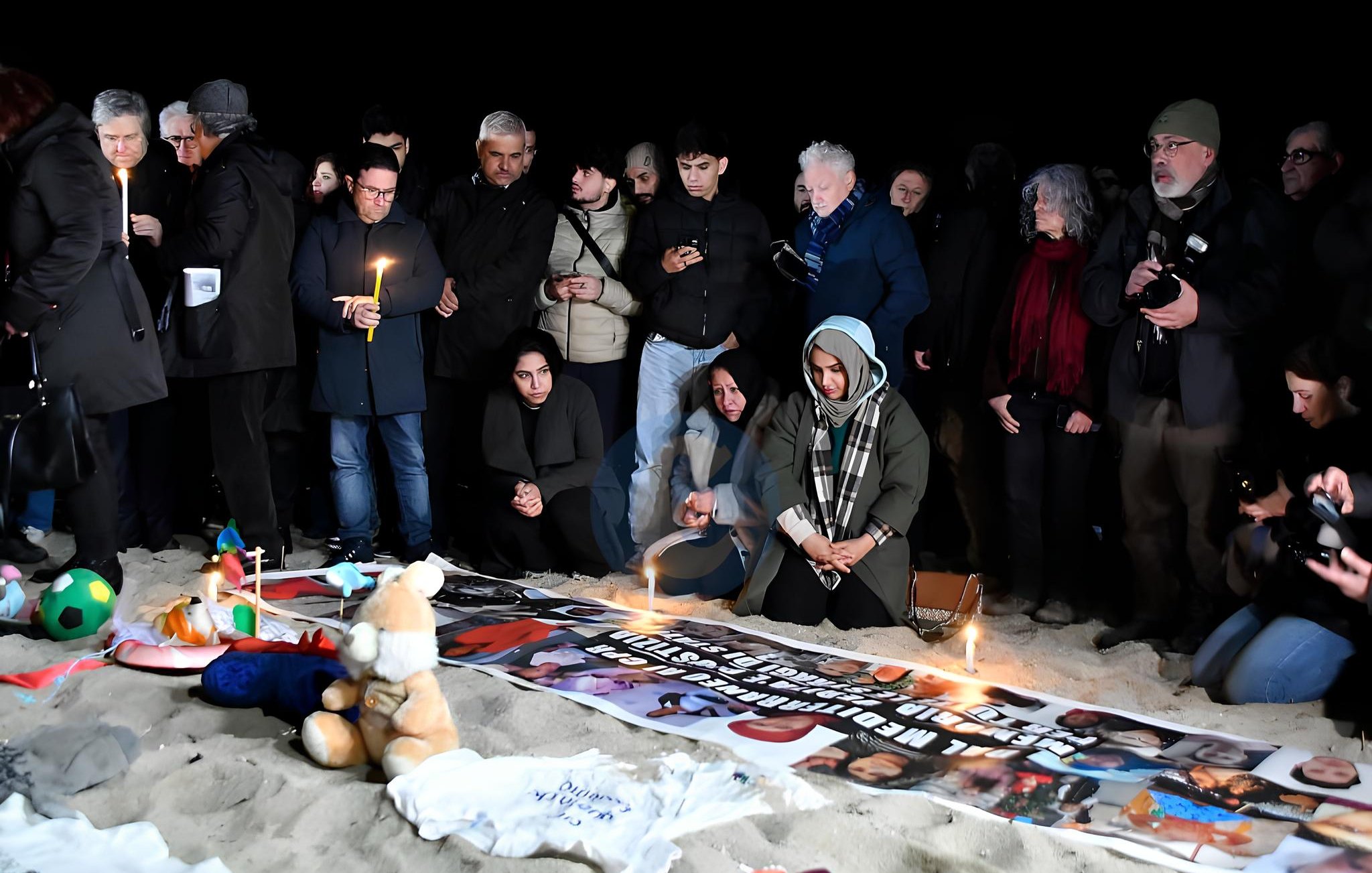 Un gruppo di persone in cerchio, con candele accese e striscioni, su una spiaggia al buio.