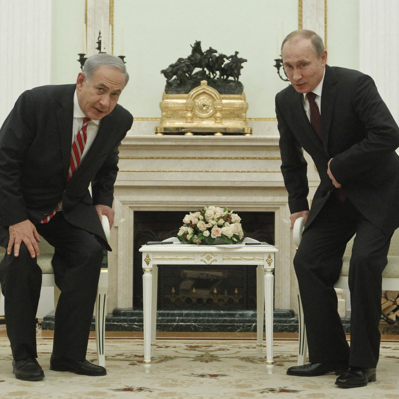 L'image montre deux hommes en costume, l'un avec une cravate rayée et l'autre avec une cravate sombre, se tenant debout devant une table basse dans une pièce élégante. Ils semblent se pencher légèrement en avant, probablement lors d'une conversation. En arrière-plan, on peut voir des éléments de décoration intérieure, dont une grande horloge et un bouquet de fleurs sur la table. Les murs sont peints dans des tons clairs et l'ensemble donne une impression de formalité.
