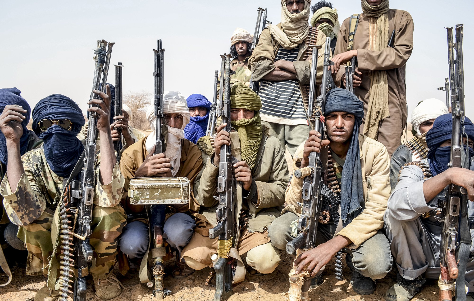 L'image montre un groupe d'hommes armés, probablement dans un environnement désertique. Ils portent des vêtements typiques de régions arides, comme des turbans et des foulards qui cachent partiellement leur visage. Chaque homme tient une arme à feu, et l'ambiance générale semble sérieuse et déterminée. Le groupe est organisé et se présente en ligne, ce qui suggère une certaine discipline. Le fond désertique renforce la dureté du cadre dans lequel ils se trouvent.