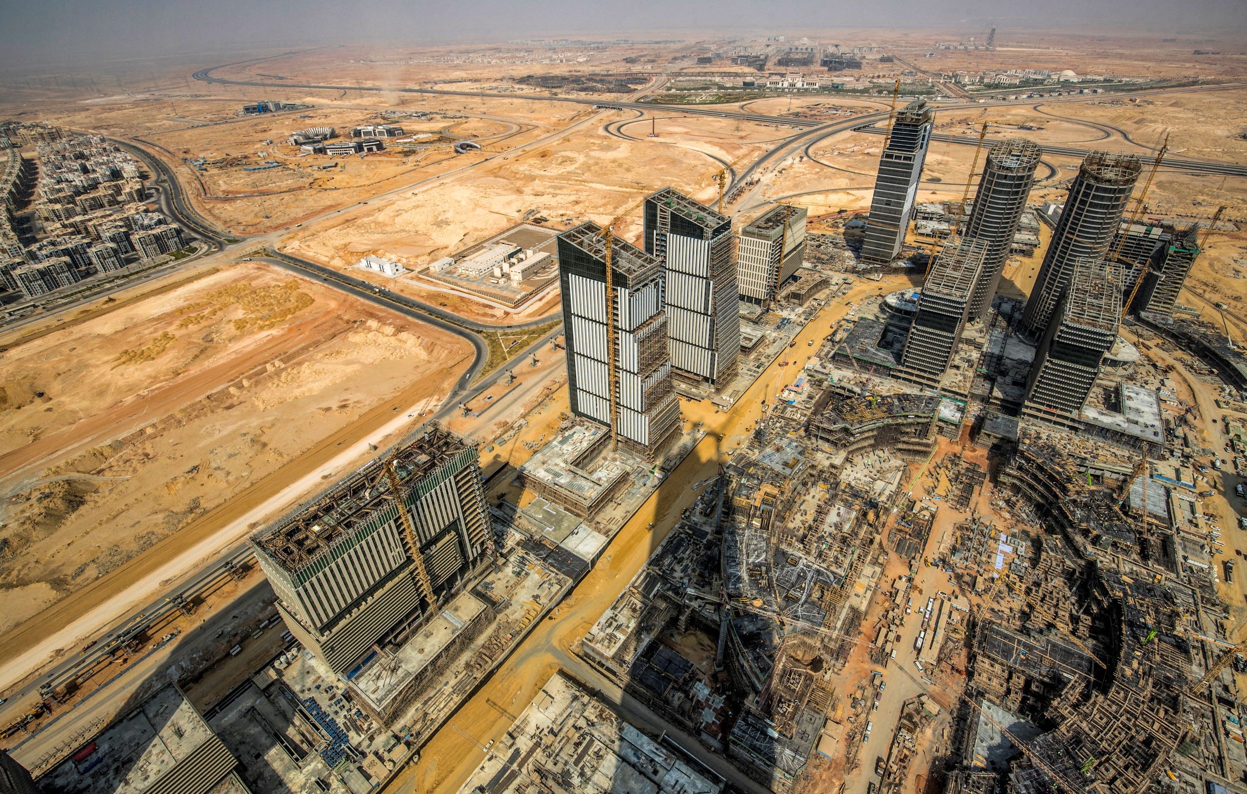The image showcases a panoramic view of a vast construction site in a desert landscape. In the foreground, several high-rise buildings are partially completed, with cranes and construction equipment evident. The surrounding area features empty land and some roadways, characteristic of a developing urban environment. The backdrop includes a clear sky and distant structures, suggesting a mixture of ongoing urbanization and natural desert terrain. The overall scene illustrates the rapid expansion of urban infrastructure in a typically arid region.