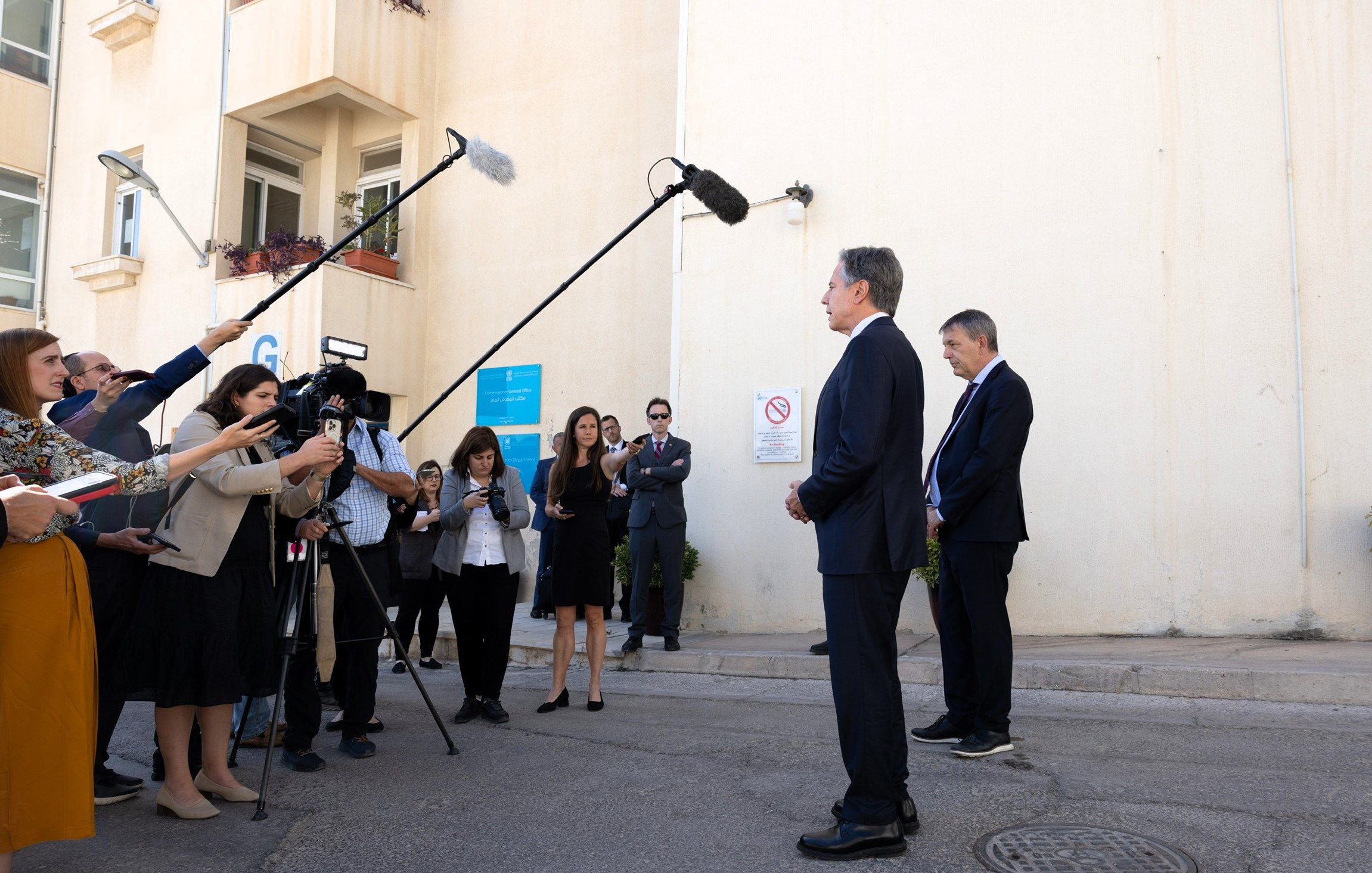 Nell'immagine si può vedere una scena di una conferenza stampa all'aperto. Due persone, vestite in abiti eleganti, sono in piedi di fronte a un muro chiaro, mentre un gruppo di giornalisti le circonda. Alcuni giornalisti stanno utilizzando microfoni e telecamere per registrare o fare domande. Dietro di loro, ci sono alcuni cartelli e avvisi su un edificio. L'atmosfera sembra formale e professionale, con un focus sulle dichiarazioni dei due individui in primo piano.