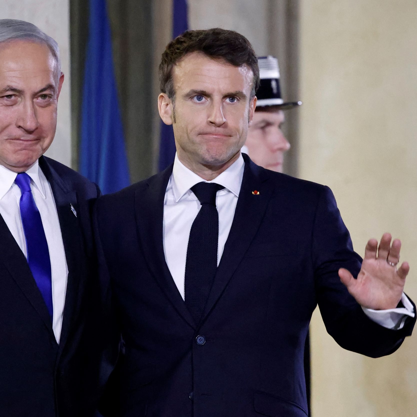 در این تصویر، دو نفر در یک موقعیت رسمی و با لبخند در کنار یکدیگر ایستاده‌اند. یکی از آن‌ها کت و کراوات تیره‌ای به تن دارد و دیگری نیز لباسی مشابه بر تن دارد. هر دو نفر به سمت دوربین نگاه می‌کنند و دستان خود را به حالت خوش‌آمد گویی بالا برده‌اند. پس‌زمینه تصویر ممکن است شامل نمادها یا پرچم‌های کشورها باشد. این تصویر به نظر می‌رسد که در یک رویداد رسمی یا کنفرانس گرفته شده است.