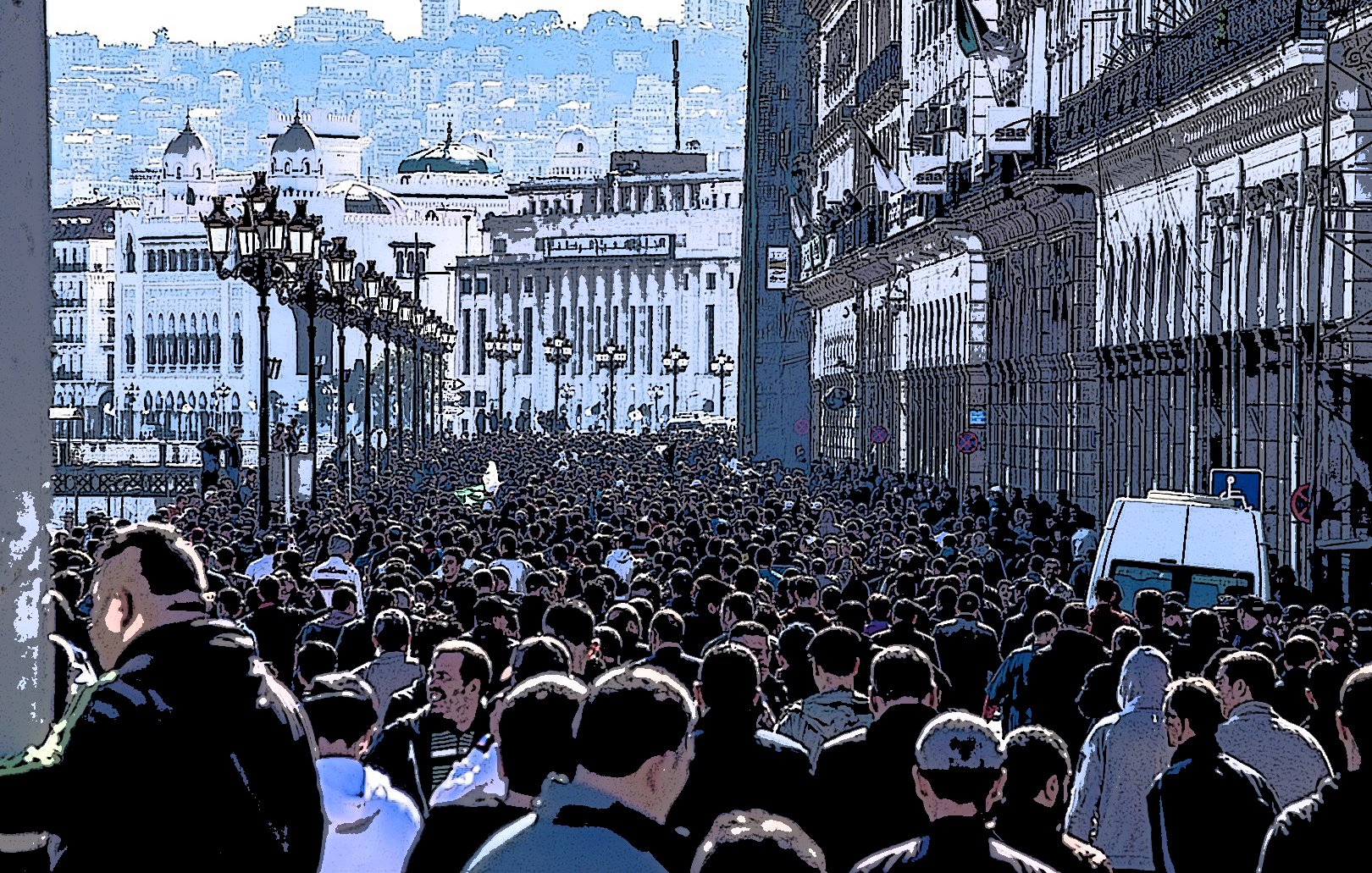 L'image montre une foule dense de personnes marchant dans une rue, probablement dans une ville animée. Les bâtiments sur les côtés semblent historiques, avec une architecture riche. On peut voir des lampadaires le long de la promenade, et au loin, on aperçoit des collines ou des montagnes qui ajoutent à la profondeur de la scène. L'atmosphère semble vibrante et dynamique, reflétant probablement une manifestation ou un événement public.