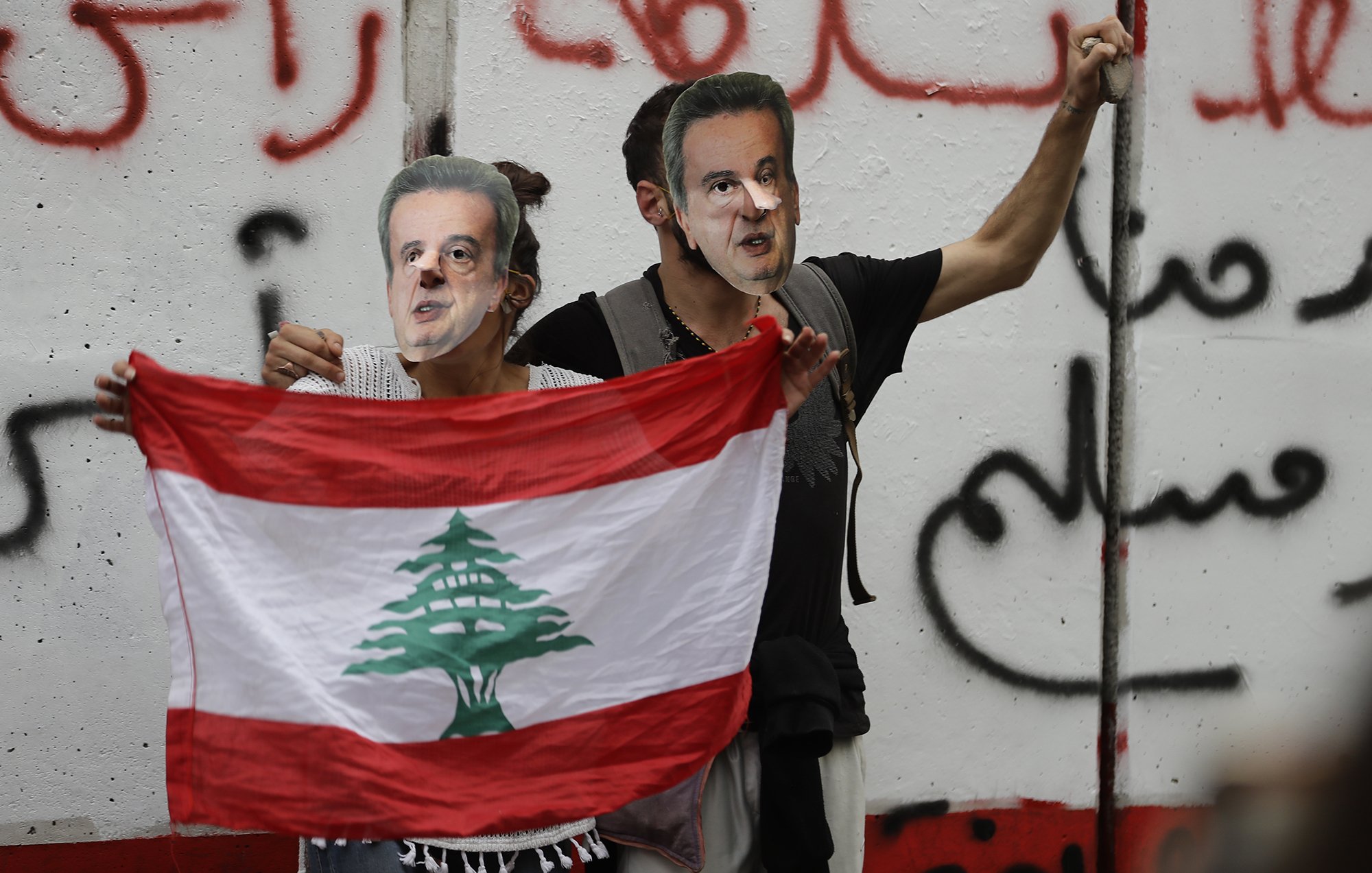 L'image montre deux personnes tenant un drapeau libanais tout en portant des masques représentant un homme. Ils se tiennent devant un mur avec des inscriptions en arabe, probablement dans un contexte de manifestation ou de protestation. L'ambiance semble chargée d'émotion et de revendications politiques.