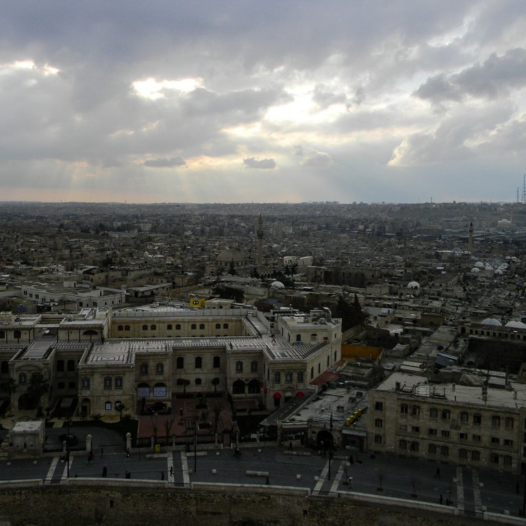 این تصویر نمای وسیعی از یک شهر را نشان می‌دهد. آسمان ابری است و پرتوهای نور از میان ابرها به زمین می‌تابد. در پیش‌زمینه، ساختمان‌های قدیمی با سقف‌های مسطح و طراحی‌های سنتی دیده می‌شوند. خیابان‌هایی در میان این ساختمان‌ها وجود دارد و در دوردست، بخشی از شهر به‌وضوح قابل مشاهده است. این نما نشان‌دهنده یک شهر تاریخی با بافتی متراکم و پیچیده است.