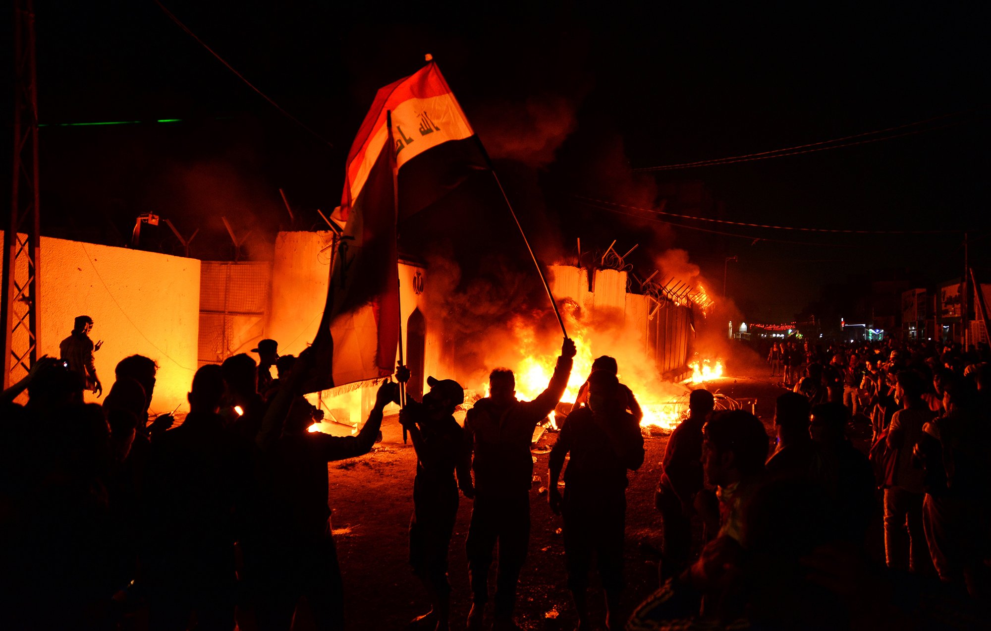 در این تصویر، گروهی از مردم در بامداد، در حال برپایی تظاهرات یا اعتراض هستند. آن‌ها در دست‌ها پرچم عراق را بلند کرده‌اند و در پس‌زمینه، آتش‌سوزی‌هایی در حال ایجاد است که موجب ایجاد نور و دود در فضا شده است. صحنه جوش و خروش و هیجان را نشان می‌دهد و احساسات قوی معترضان را به تصویر می‌کشد. دیوارهایی در اطراف دیده می‌شود که ممكن است بخشی از یک ساختمان یا منطقه محصور باشد.