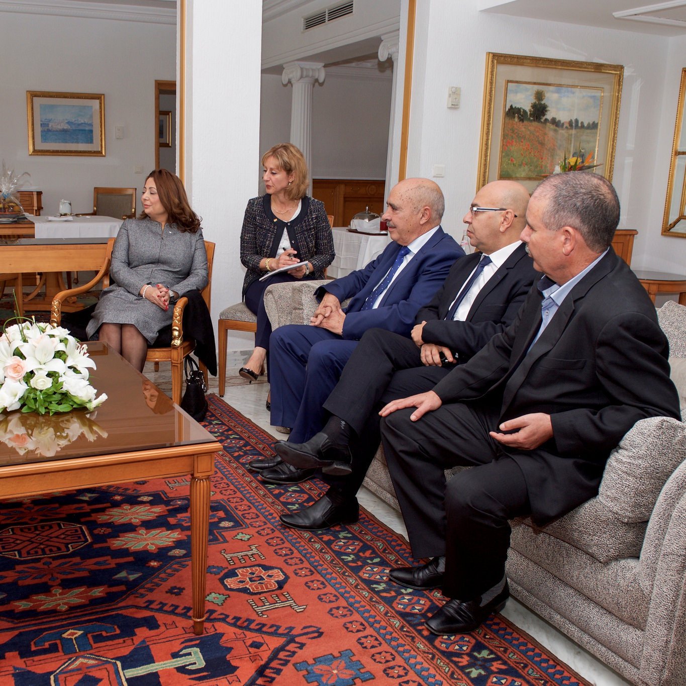 L'image montre un groupe de personnes assises dans un salon bien éclairé et décoré. Il y a une table basse au centre avec un arrangement floral blanc. À gauche, une personne est assise dans un fauteuil, écoutant attentivement. Les autres personnes sont disposées autour de la table, portant des costumes formels et engageant une conversation. Les murs sont ornés de tableaux, et le décor évoque une atmosphère professionnelle et conviviale.