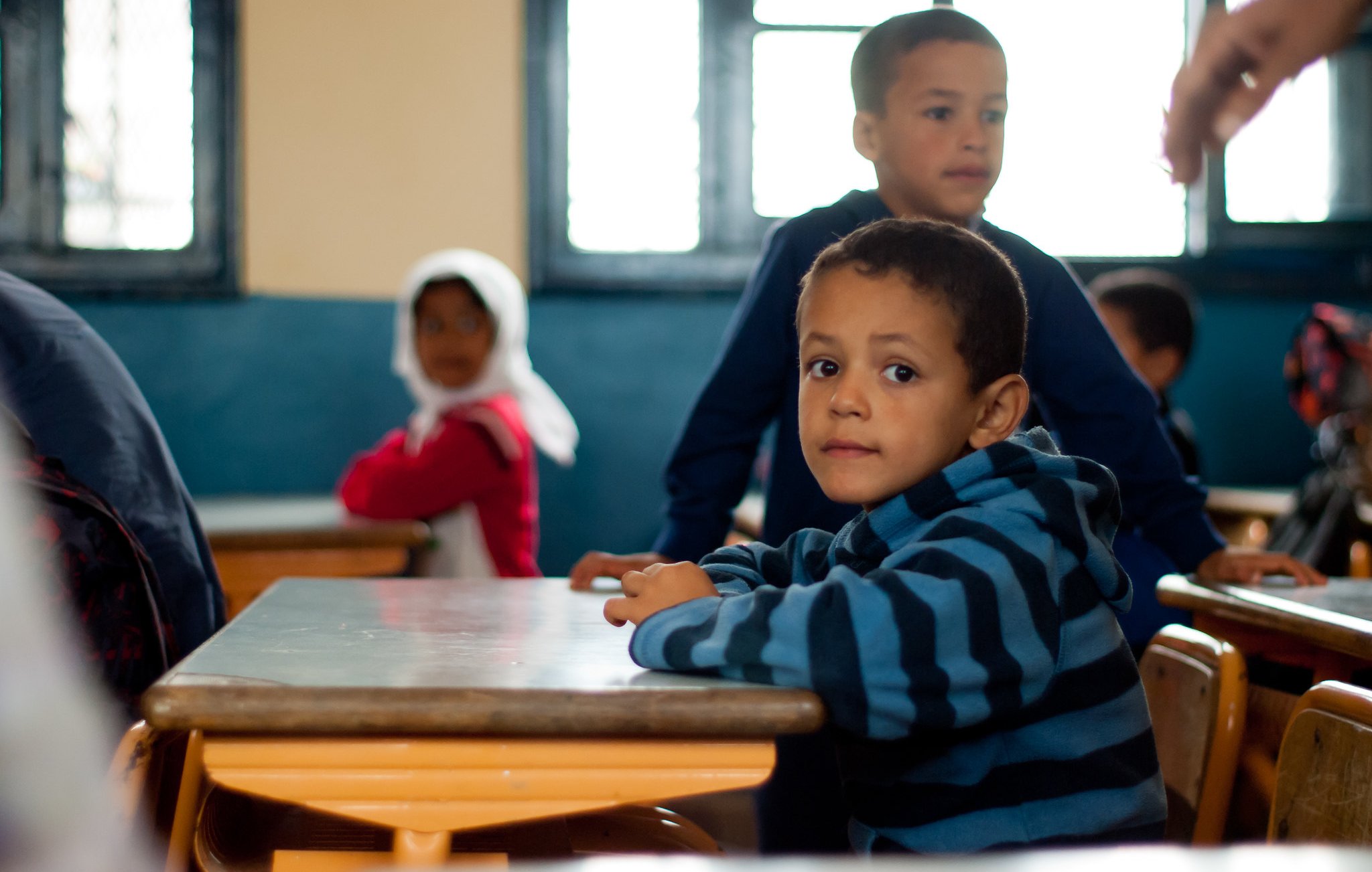 الصورة تُظهر مجموعة من الأطفال في فصل دراسي. في المقدمة، يوجد طفل يجلس على الطاولة، يرتدي سترة مخططة بألوان زرقاء وخضراء، يبدو عليه الفضول وهو ينظر نحو الكاميرا. خلفه، يوجد طفل آخر واقف، بالإضافة إلى فتاة ترتدي حجابًا وتجلس في الخلف. الأجواء توحي بالعلم والتعلم، مع وجود النوافذ التي تسمح بدخول الضوء.