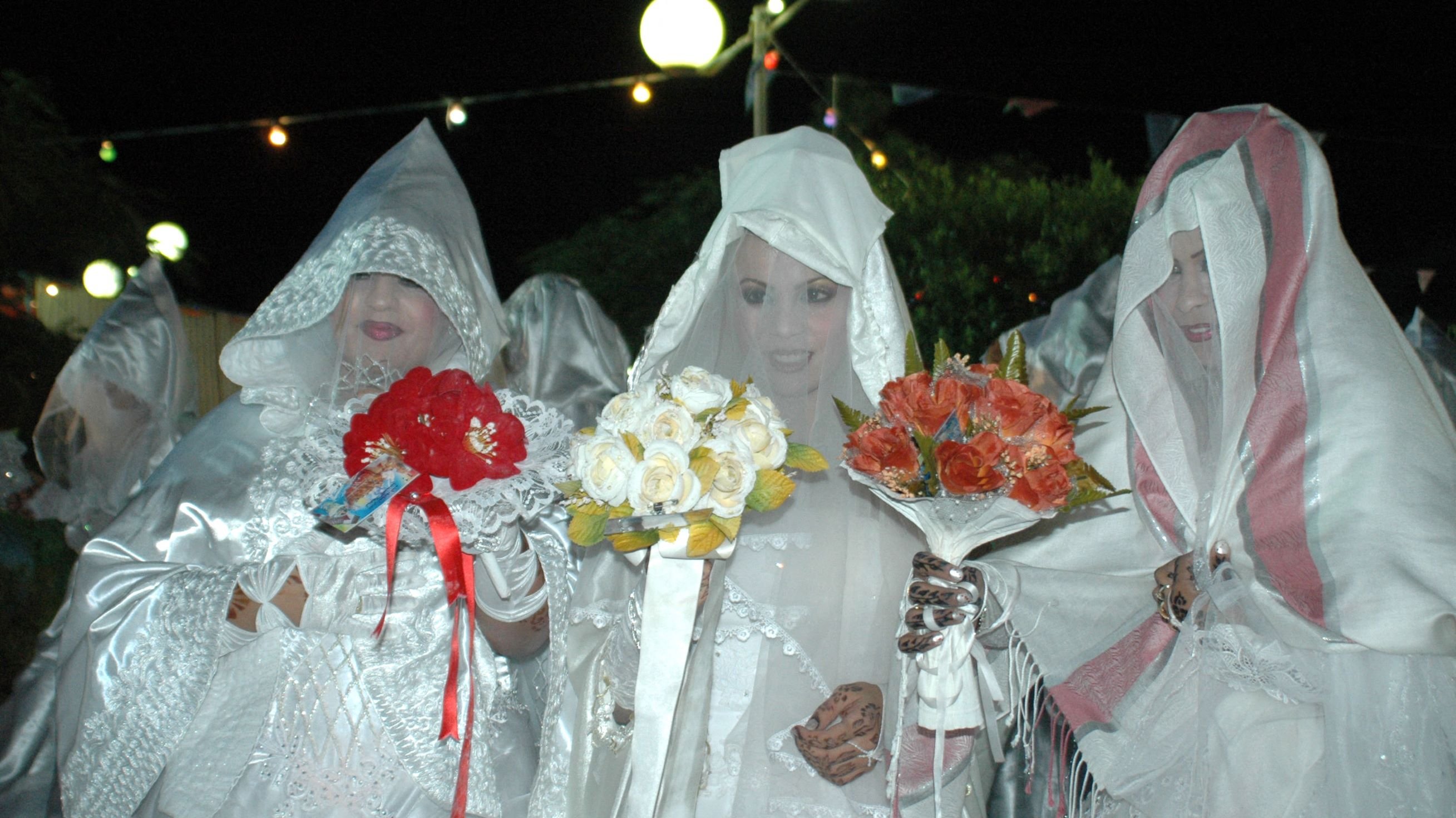 L'immagine mostra un gruppo di donne vestite con abiti bianchi e coperti da veli, che formano un'atmosfera suggestiva. Alcune di loro tengono in mano mazzi di fiori colorati, come rose rosse e fiori bianchi. L'illuminazione è soffusa, creando un'atmosfera notturna, e sullo sfondo si possono vedere luci decorative. Le donne sembrano partecipare a un evento particolare, probabilmente una celebrazione o un rituale tradizionale.