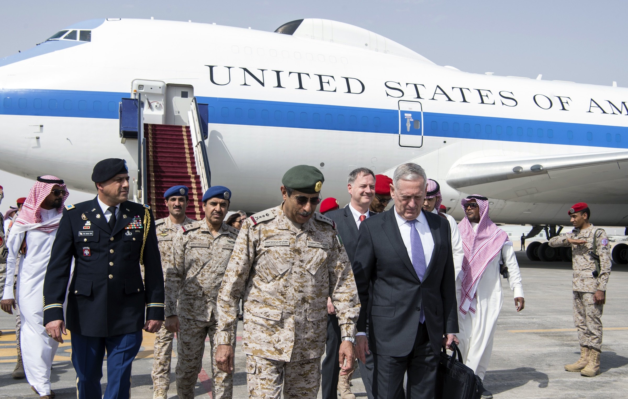 The image depicts a group of individuals exiting from a large aircraft marked with the words "UNITED STATES OF AMERICA." In the foreground, there are several military personnel and officials. One individual appears to be dressed in a military uniform, while others are in formal attire. They seem to be engaged in conversation as they walk down the steps from the plane. The backdrop includes the aircraft and an airport setting, suggesting a diplomatic visit or military engagement.