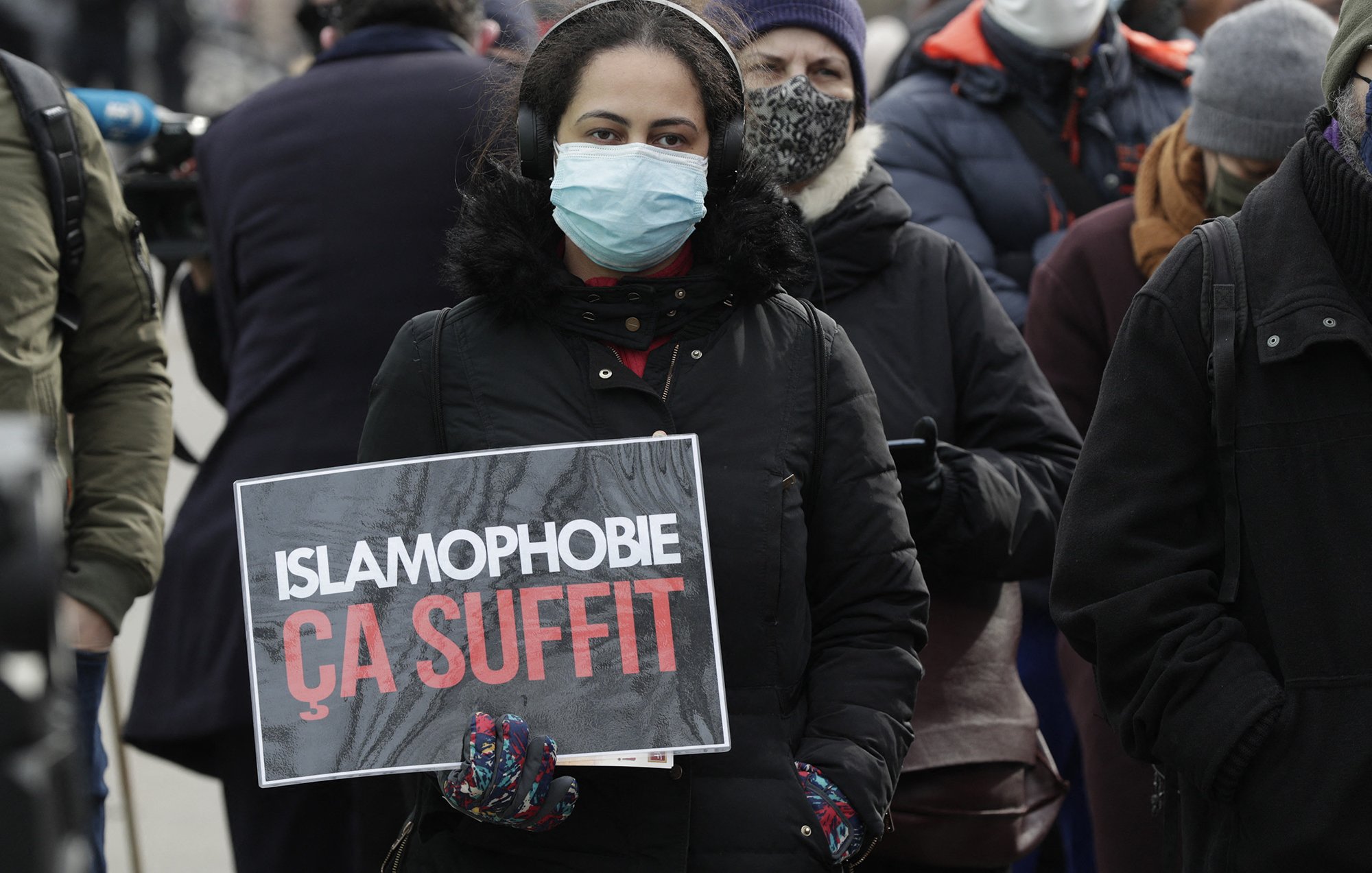 En la imagen se puede ver a una persona sosteniendo un cartel que dice "ISLAMOPHOBIE ÇA SUFFIT", lo que se traduce como "ISLAMOFOBIA BASTA YA". La persona lleva puesta una mascarilla y parece estar en un evento o manifestación, rodeada de otras personas que también usan mascarillas. El ambiente sugiere una expresión de protesta o de apoyo a una causa relacionada con la lucha contra la islamofobia.