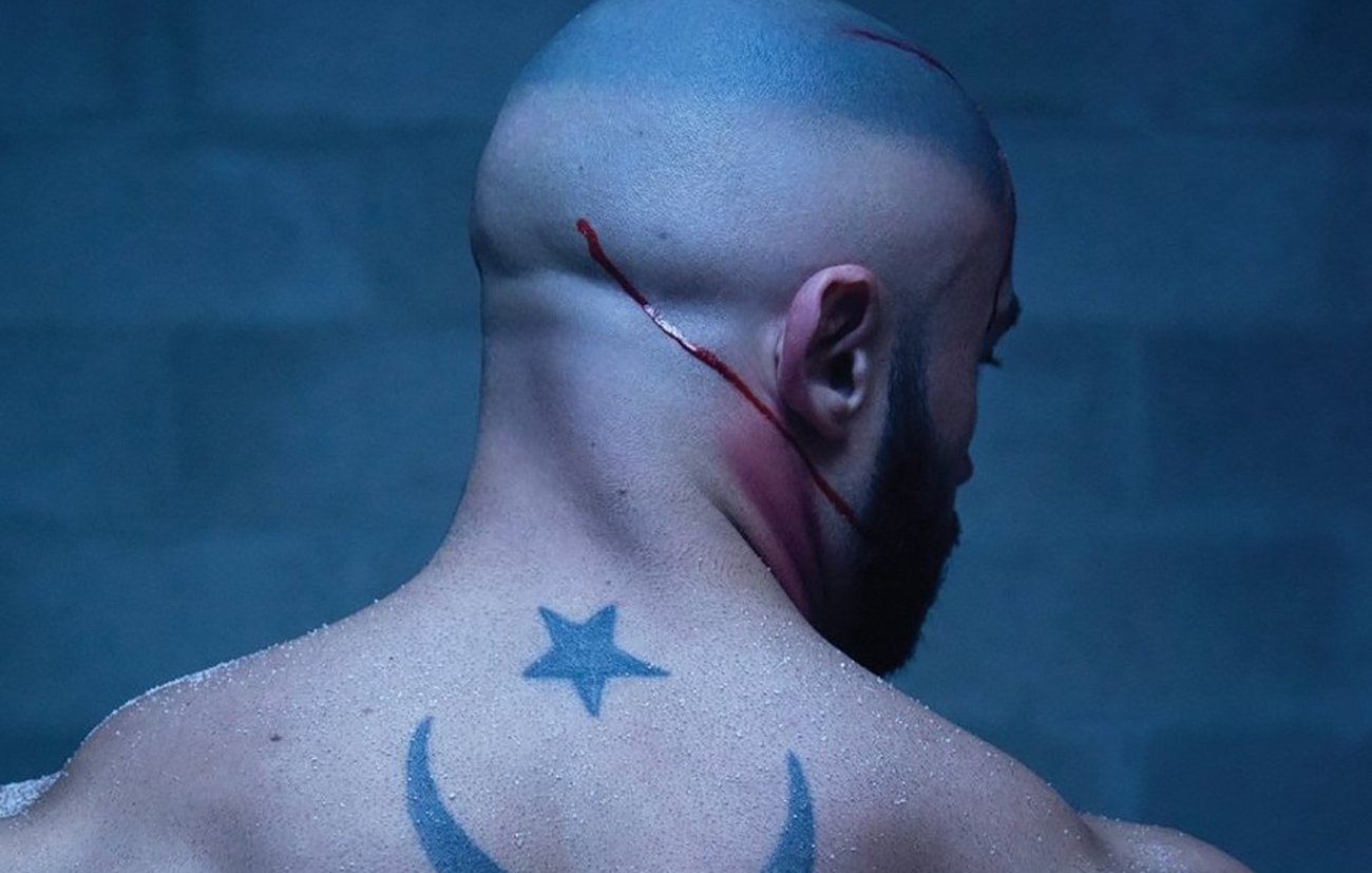 The image depicts a muscular, bald man from the back, showcasing a prominent tattoo on his back. The tattoo features a crescent moon and a star, which are often associated with various cultural and religious symbols. There are visible scars or cuts on his scalp and a streak of blood running down, suggesting a recent injury or intense situation. The background has a dark, moody atmosphere, possibly indicating a gritty or dramatic context.
