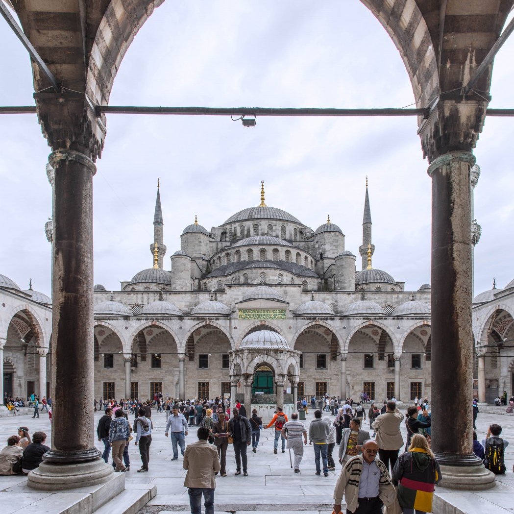 The image features a grand architectural view of the Sultan Ahmed Mosque, commonly known as the Blue Mosque, in Istanbul, Turkey. In the foreground, you can see a bustling courtyard filled with visitors and tourists. The mosque's impressive structure is visible in the background, characterized by its large dome and multiple tall minarets reaching upward. The scene is framed by arched columns, adding to the architectural beauty. The sky appears overcast, creating a soft light that enhances the details of the mosque's intricate design.