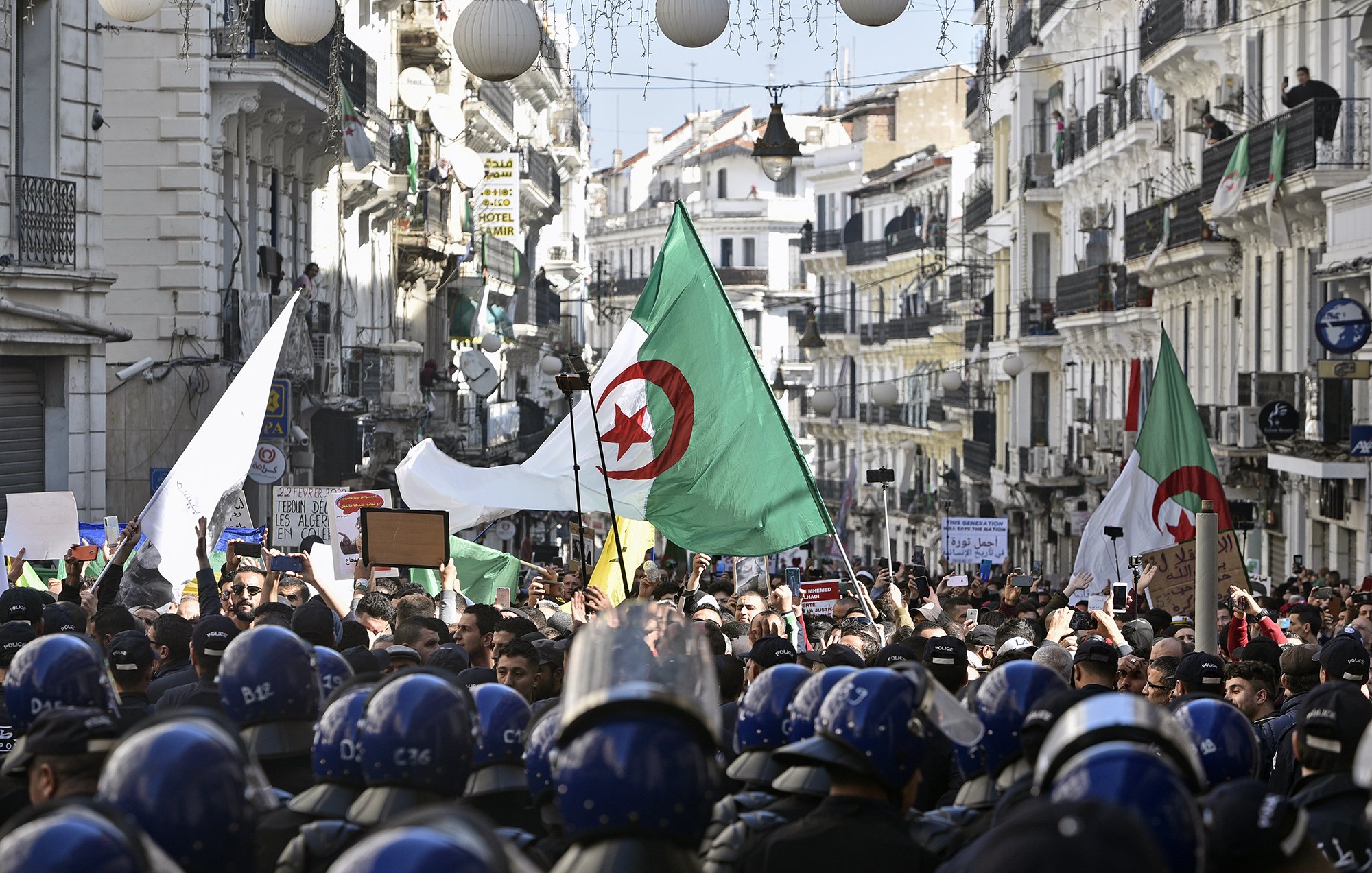 L'image montre une foule rassemblée dans une rue animée, probablement dans une ville algérienne, en période de manifestation. Des personnes portent des drapeaux algériens et tiennent des pancartes, tandis qu'un groupe de policiers en uniforme, portant des casques, se tient en ligne devant eux. Les bâtiments alentours sont typiques d'une architecture urbaine, ornés de décorations. L'atmosphère semble être celle d'une démonstration populaire, avec un fort engagement civique visible parmi les manifestants.