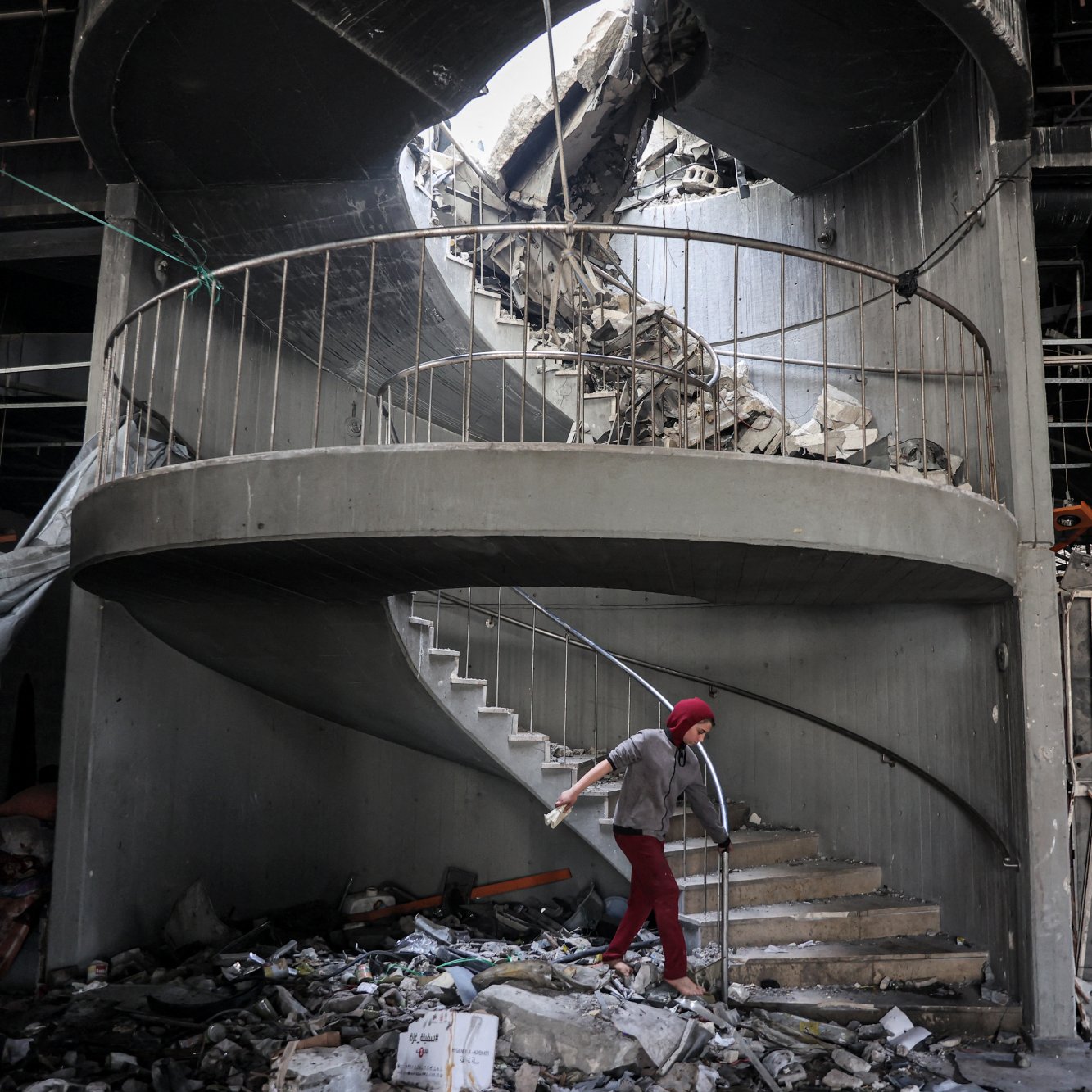 در این تصویر، یک پله‌گردان دیده می‌شود که به دلیل تخریب، وضعیت نامناسبی دارد. بخشی از سقف بالای پله‌ها و دیوارها فرو ریخته و آوار زیادی در اطراف پله‌ها جمع شده است. یک فرد با عصا در حال حرکت در پله‌ها است و به نظر می‌رسد در تلاش است تا از این فضای خراب عبور کند. نور طبیعی از سقف خراب به داخل تابیده و فضایی دلگیر و غم‌انگیز به وجود آورده است.