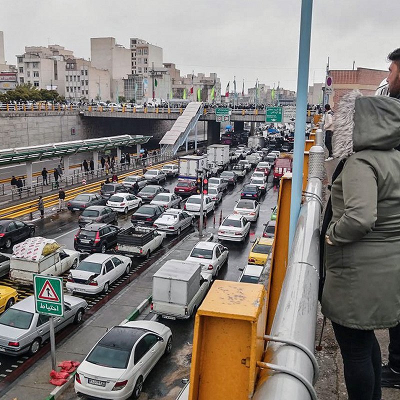 تصویر نشان‌دهنده یک صحنه شلوغ از ترافیک در یک خیابان شهری است. در سمت چپ تصویر، افرادی در حال تماشا از بالا ایستاده‌اند. در پایین، خطوط خودروها به وضوح قابل مشاهده است که نشان‌دهنده تجمع و ترافیک سنگین است. بقیه عناصر شهری مانند ساختمان‌ها و علائم راهنمایی نیز در پس‌زمینه دیده می‌شوند. هوای ابری و احتمالا بارانی به دید فضا و حس و حال تصویر اضافه می‌کند.