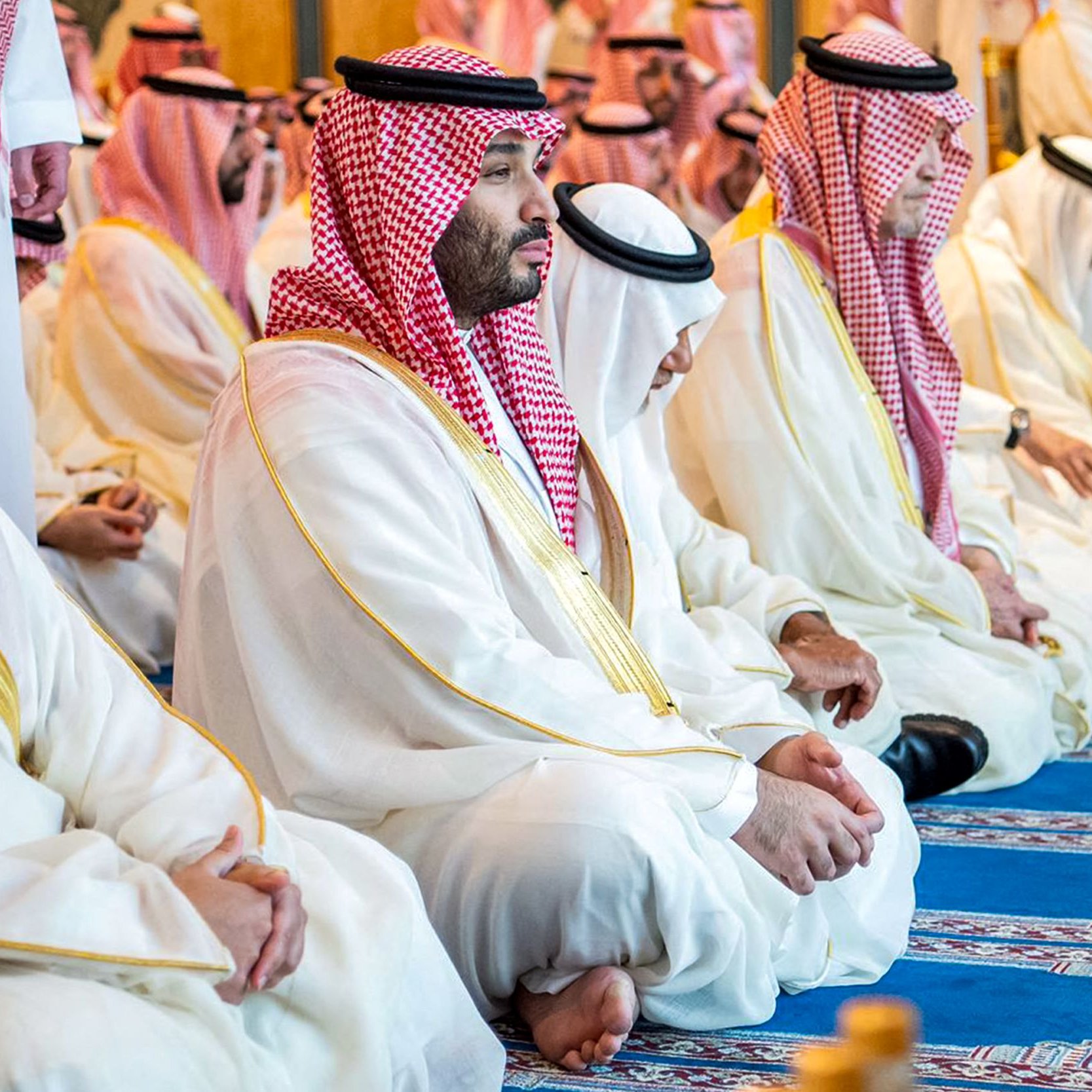 این تصویر گروهی از مردان را در حال نشسته و دعا در یک مکان مذهبی نشان می‌دهد. همه آن‌ها لباس‌های سنتی سعودی، شامل عبا و گتر (کلاه عربی) به تن دارند. بعضی از آن‌ها به طور جدی و متمرکز در حال دعا هستند و فضای کلی تصویر حالتی معنوی و آرام دارد. زمینه تصویر نیز شامل فرش و دیوارهایی با طراحی‌های داخلی شیک است.