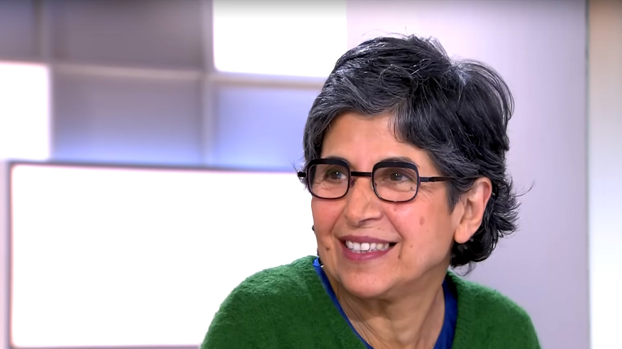 L'image montre une femme souriante avec des cheveux gris et courts. Elle porte des lunettes et un pull de couleur verte. Son expression est amicale et chaleureuse, indiquant qu'elle participe à une conversation. L'arrière-plan est flou, suggérant qu'elle se trouve dans un environnement de studio ou d'interview.