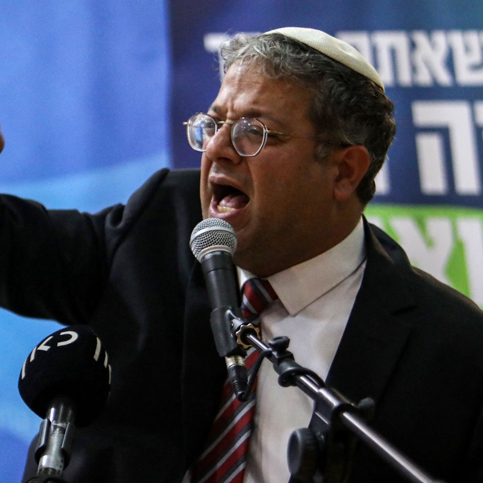 تظهر الصورة شخصية تتحدث بحماسة أمام ميكروفون خلال مؤتمر أو حدث عام. يرتدي بدلة رسمية ونظارات، ويبدو أنه يعبر عن مشاعر قوية، حيث يشير بيده ويظهر تعابير وجه تعكس الحماس أو الغضب. الخلفية تحتوي على شعارات أو نصوص، مما يدل على أن الحدث قد يكون سياسيًا أو يتعلق بقضية عامة.