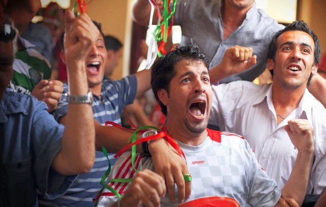 L'image montre un groupe de jeunes hommes dans une ambiance festive. Ils semblent très joyeux et enthousiastes, certains criant et souriant, probablement en train de célébrer un événement sportif. Ils portent des vêtements décontractés et il y a des décorations colorées autour d'eux. Au premier plan, un homme exprime sa joie de manière très expressive, avec les bras levés. En bas, on peut voir un slogan qui dit "LA VIE VOUS APPELLE". L'ensemble de la scène dégage une atmosphère de camaraderie et de bonheur collectif.