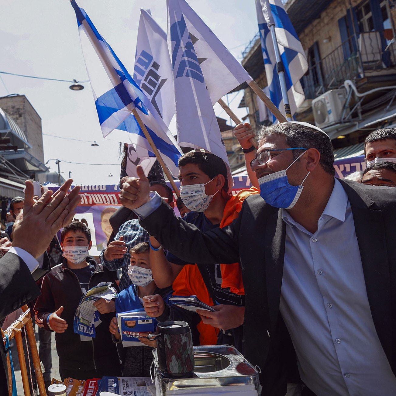 در این تصویر، مردی با ریش بلند و کلاه سیاه در حال سخنرانی و برقراری تعامل با جمعیتی است که پرچم‌های اسرائیل را در دست دارند. افراد حاضر در تصویر، که تعدادی از آن‌ها ماسک به صورت دارند، به نظر می‌رسد در حال ابراز حمایت یا اعتراض هستند. فضا به شدت شلوغ و پرانرژی به نظر می‌رسد و به وضوح نشان‌دهنده یک رویداد اجتماعی یا سیاسی است. ساختارها و تجسس‌های شهری نیز در پس‌زمینه دیده می‌شود.