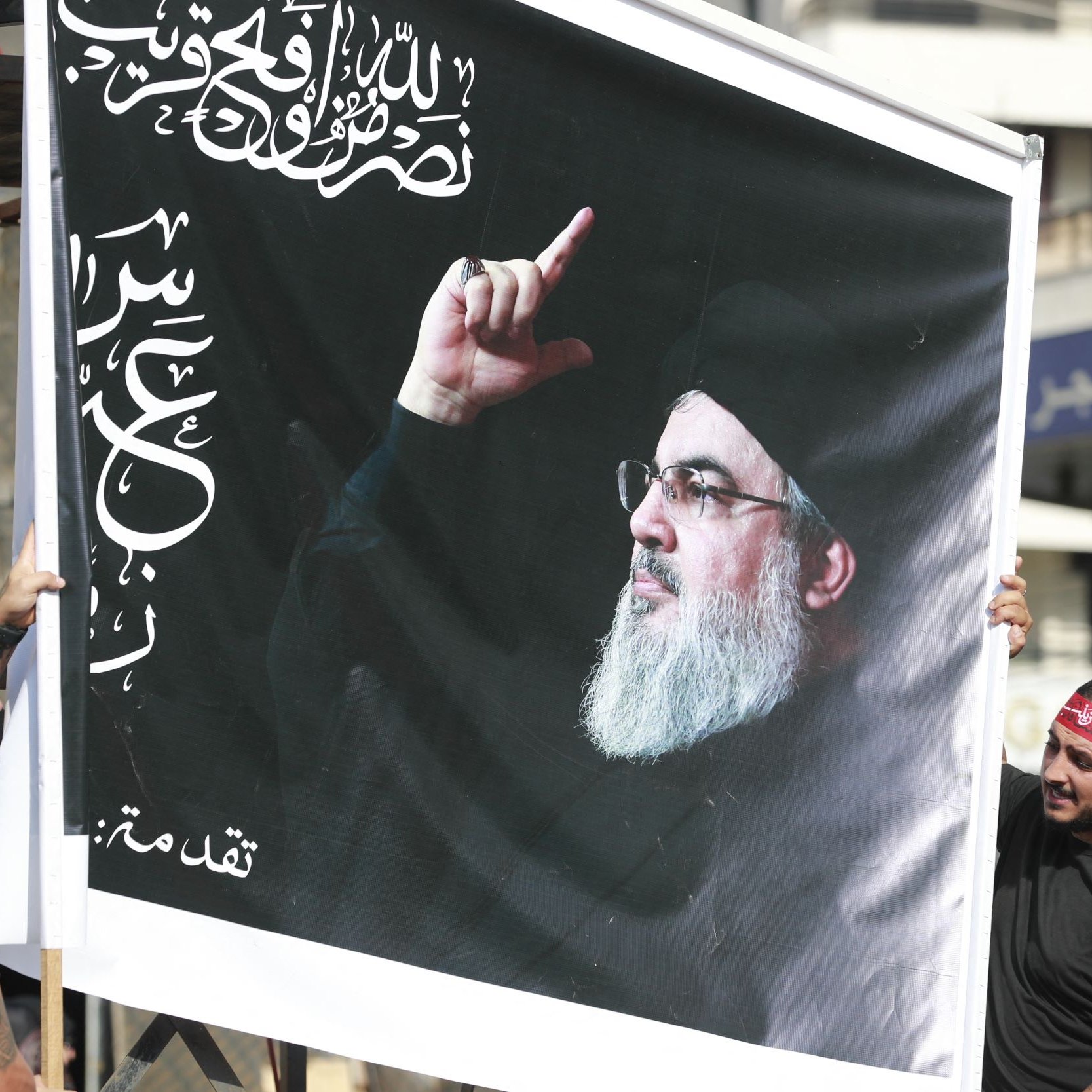 L'image montre deux personnes tenant une grande affiche. Cette affiche représente un homme avec une barbe, portant un vêtement sombre, et levant un doigt. En arrière-plan, on aperçoit des bâtiments. Les deux personnes portent des bandanas rouges et des t-shirts noirs. L'affiche comporte également des inscriptions arabes.