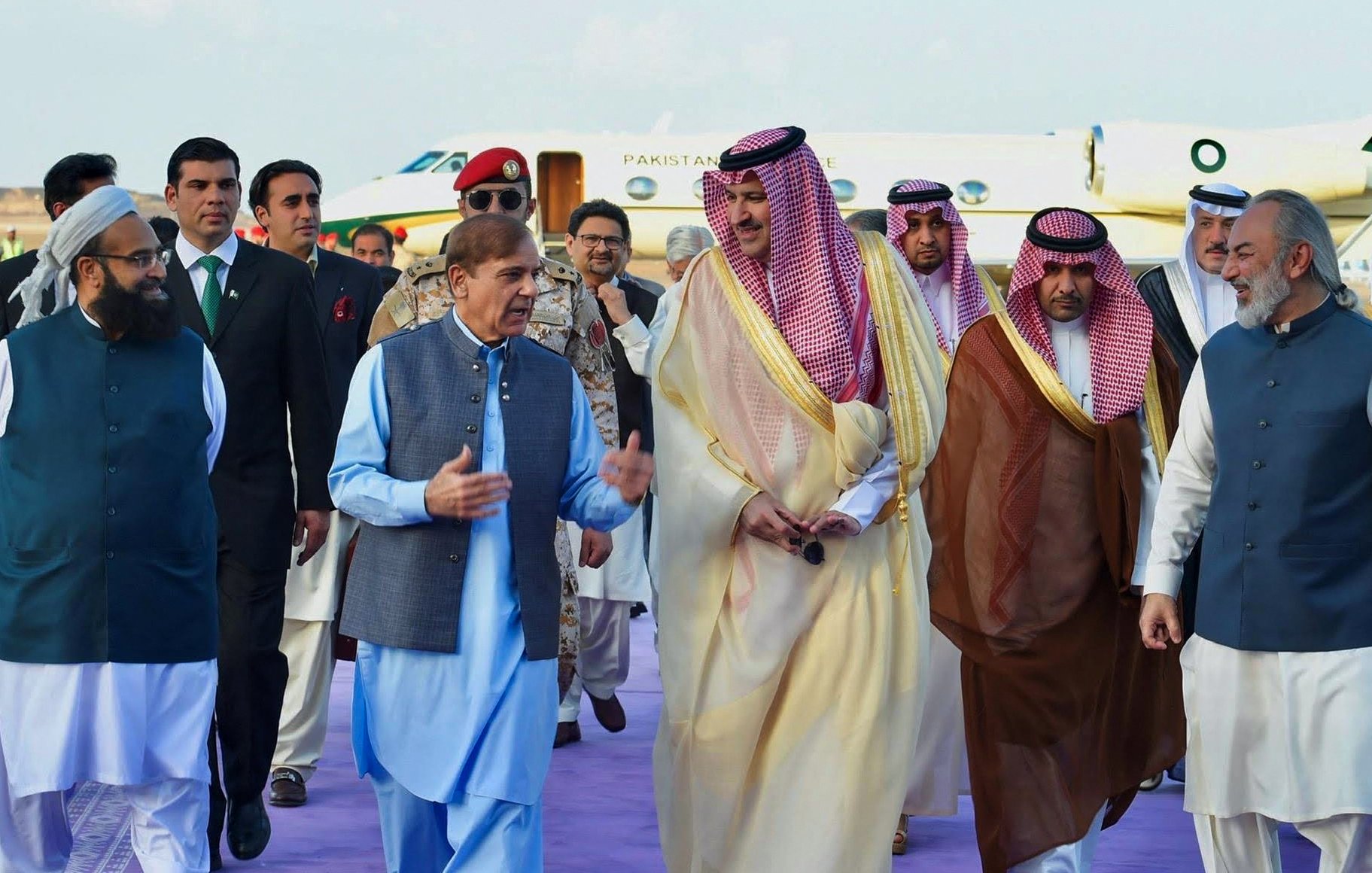 في الصورة، يظهر عدد من الشخصيات في احتفال أو مناسبة رسمية. يقود المجموعة شخص يرتدي زيًا تقليديًا باللون الأزرق، ويتحدث بوضوح مع الآخرين الذين يرتدون ملابس تقليدية مختلفة. يبدو أن هناك تفاعلاً ودودًا بينهم. في الخلفية، يظهر طائرة خاصة، مما يشير إلى أن الحدث قد يكون متعلقًا بالسفر أو زيارة رسمية. الأجواء تبدو احتفالية وتضم مجموعة متنوعة من الأشخاص الذين يمثلون ثقافات مختلفة.