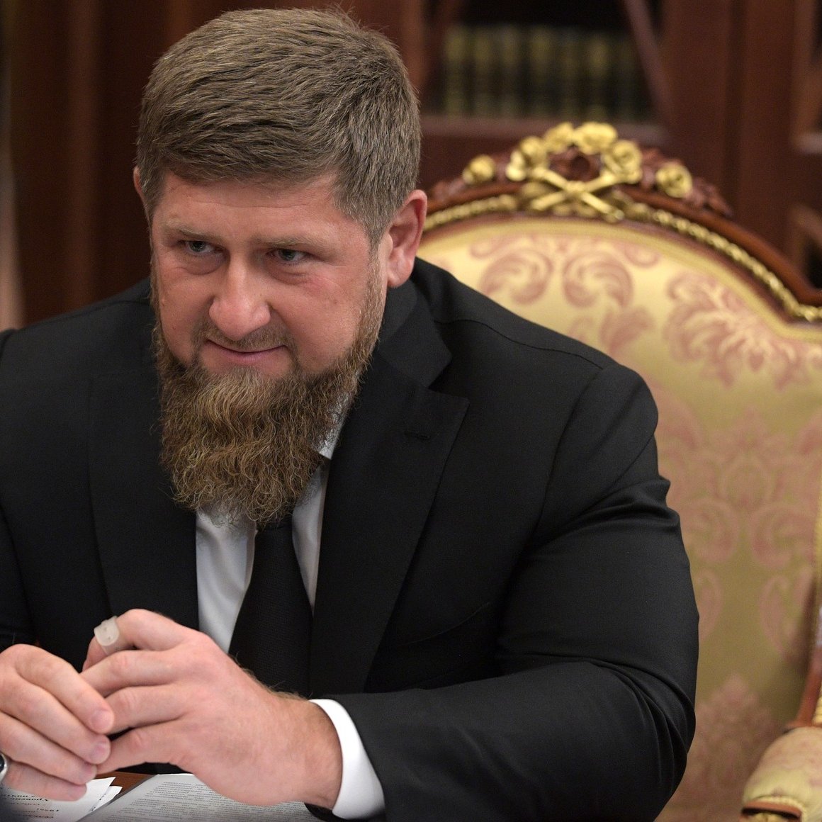 الصورة تظهر رجلًا يجلس على طاولة، وهو يرتدي بدلة رسمية سوداء. يتمتع بلحية كثيفة ويمسك بيديه شيئًا ما. خلفه تظهر تفاصيل من مكتب فاخرة، مثل الأثاث المزخرف والمكتبات. يبدو أنه في اجتماع أو مناقشة مع شخص آخر.