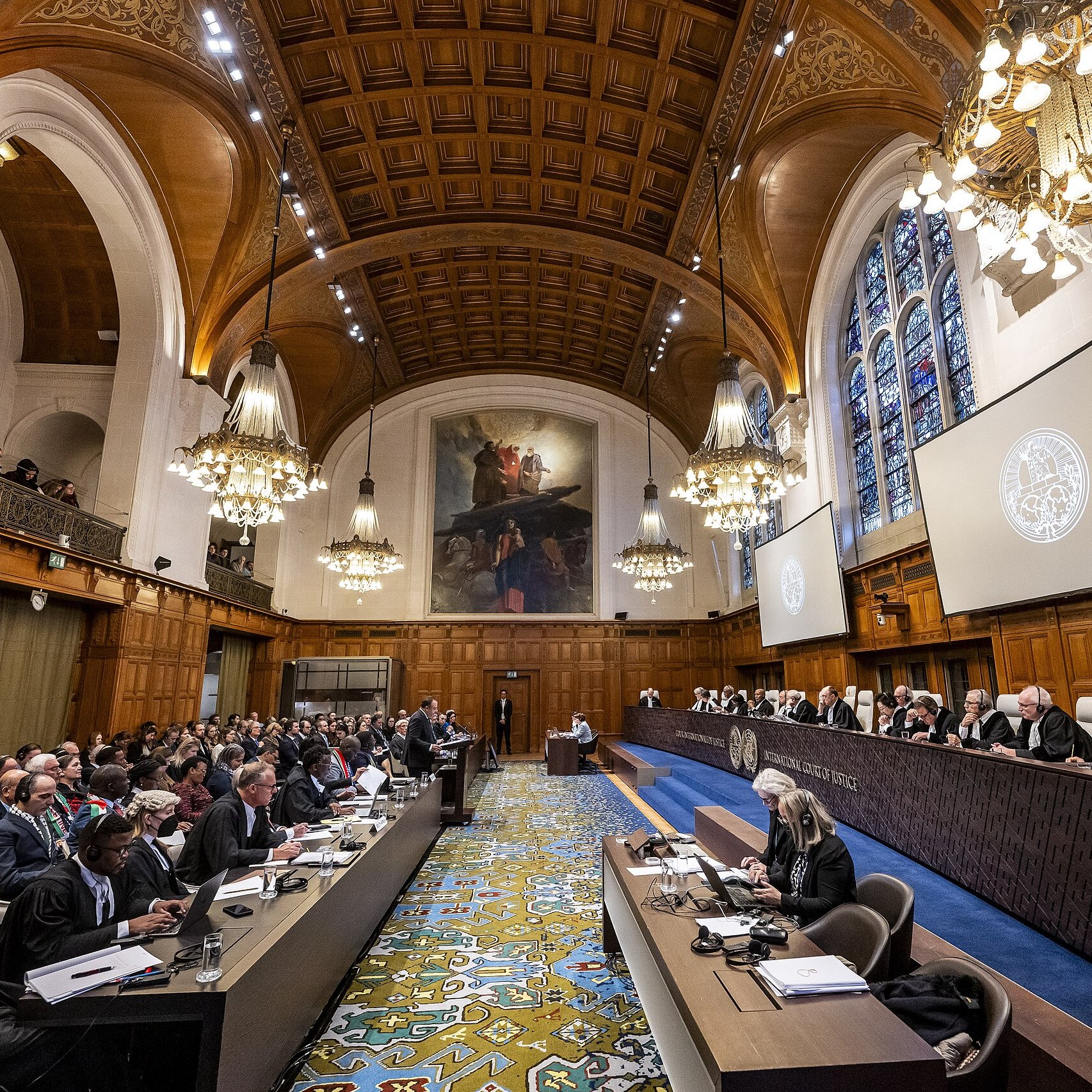 L'immagine mostra un'aula di giustizia, probabilmente una corte internazionale. La sala è caratterizzata da un'architettura imponente con soffitti alti, lampadari eleganti e finestre ad arco che lasciano filtrare la luce. In primo piano, c'è un lungo tavolo con diversi avvocati e rappresentanti, mentre sul lato opposto, una serie di giudici sono seduti su una piattaforma rialzata. Il pubblico è composto da molte persone, alcune delle quali prendono appunti. L'atmosfera sembra formale e rispettosa, tipica di un'importante udienza legale.