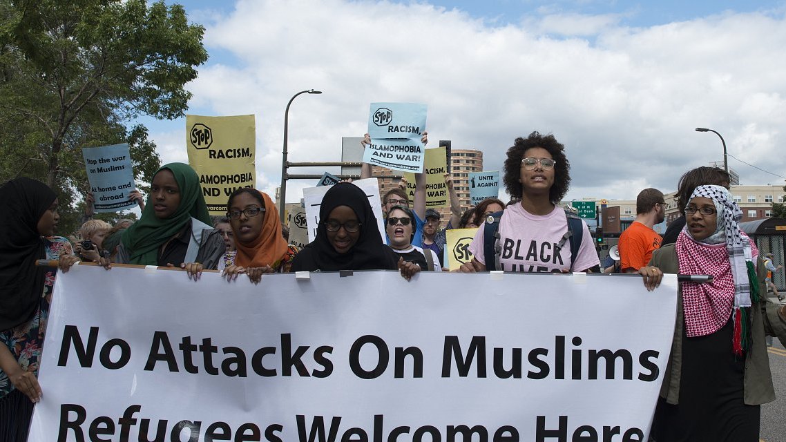 في الصورة، تظهر مجموعة من الأشخاص المشاركين في مظاهرة. يحمل المتظاهرون لافتات مكتوب عليها عبارات تدعو للتضامن مع المسلمين وترحب باللاجئين. يرتدي بعض النساء الحجاب، وتظهر تعابير الحماس والتأييد على وجوههم. السماء تبدو غائمة قليلاً، مما يضفي توتراً على الأجواء، وفي الخلفية يمكن رؤية مب buildings وشعارات تعبر عن رفض العنصرية والتمييز.