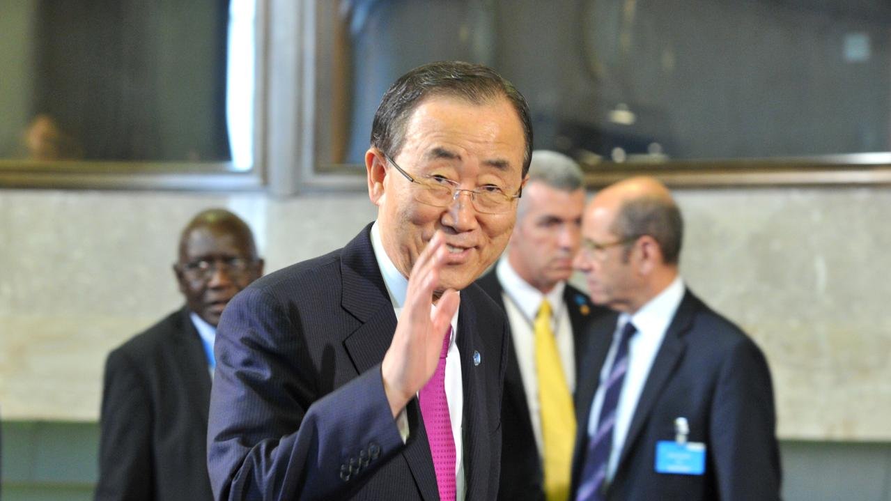 L'image montre un homme souriant, vêtu d'un costume sombre avec une cravate rose. Il semble faire un geste de la main, comme s'il saluait. En arrière-plan, on peut apercevoir d'autres hommes en costume, certains portant des badges. L'environnement semble être un lieu formel, probablement dans un contexte de réunion ou de conférence internationale.