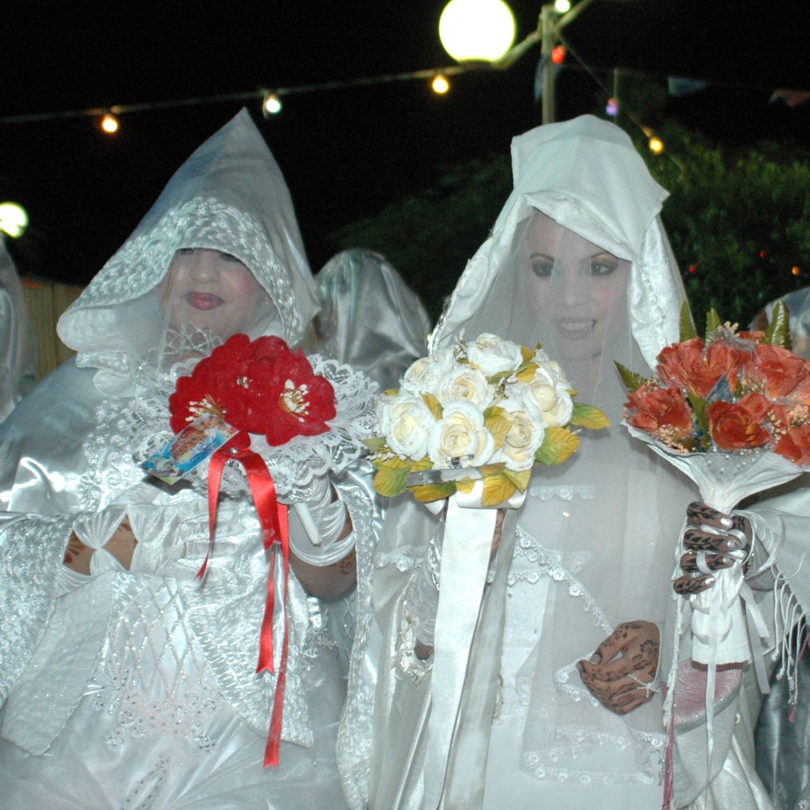 L'immagine mostra un gruppo di donne vestite con abiti bianchi e coperti da veli, che formano un'atmosfera suggestiva. Alcune di loro tengono in mano mazzi di fiori colorati, come rose rosse e fiori bianchi. L'illuminazione è soffusa, creando un'atmosfera notturna, e sullo sfondo si possono vedere luci decorative. Le donne sembrano partecipare a un evento particolare, probabilmente una celebrazione o un rituale tradizionale.