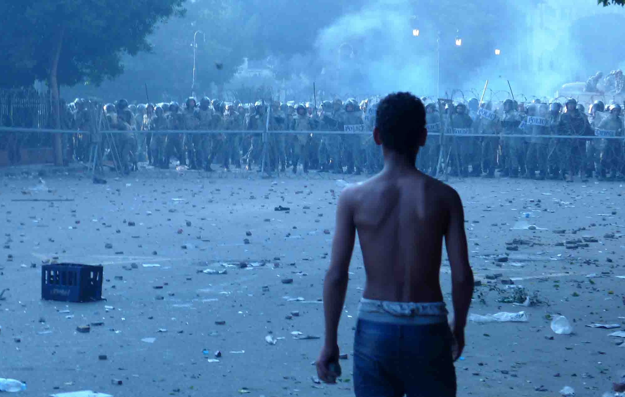 L'image montre un jeune garçon, torse nu, qui se tient de dos face à une ligne de forces de l'ordre en uniforme. Il est dans un environnement troublé, avec des débris et des bouteilles éparpillés au sol, ainsi qu'une légère brume qui donne une atmosphère tendue et chaotique. La scène semble se dérouler dans un contexte de troubles ou de manifestations. Les policiers, en arrière-plan, se tiennent en formation, suggérant un affrontement imminent.