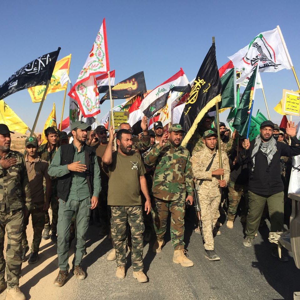 الصورة تظهر مجموعة من الأشخاص يسيرون معًا في مظاهرة أو مسيرة، يحملون أعلامًا متعددة الألوان. يبدو أنهم يرتدون ملابس عسكرية وبعضهم يرتدي زياً تقليدياً. الخلفية طبيعية، مع أراضٍ جافة وصحراوية. تعكس الصورة أجواء حماسية وتضامن بين المشاركين.