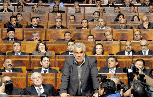 L'image montre une séance dans un bâtiment législatif, où une personne (probablement un membre du gouvernement ou un habile orateur) se tient devant un grand groupe de personnes assises. Les membres de l'assemblée, en arrière-plan, semblent concentrés sur le discours prononcé. On peut voir des journalistes avec des caméras, suggérant que cet événement est couvert par les médias. L'atmosphère est formelle et sérieuse, typique de débats ou de discussions politiques.