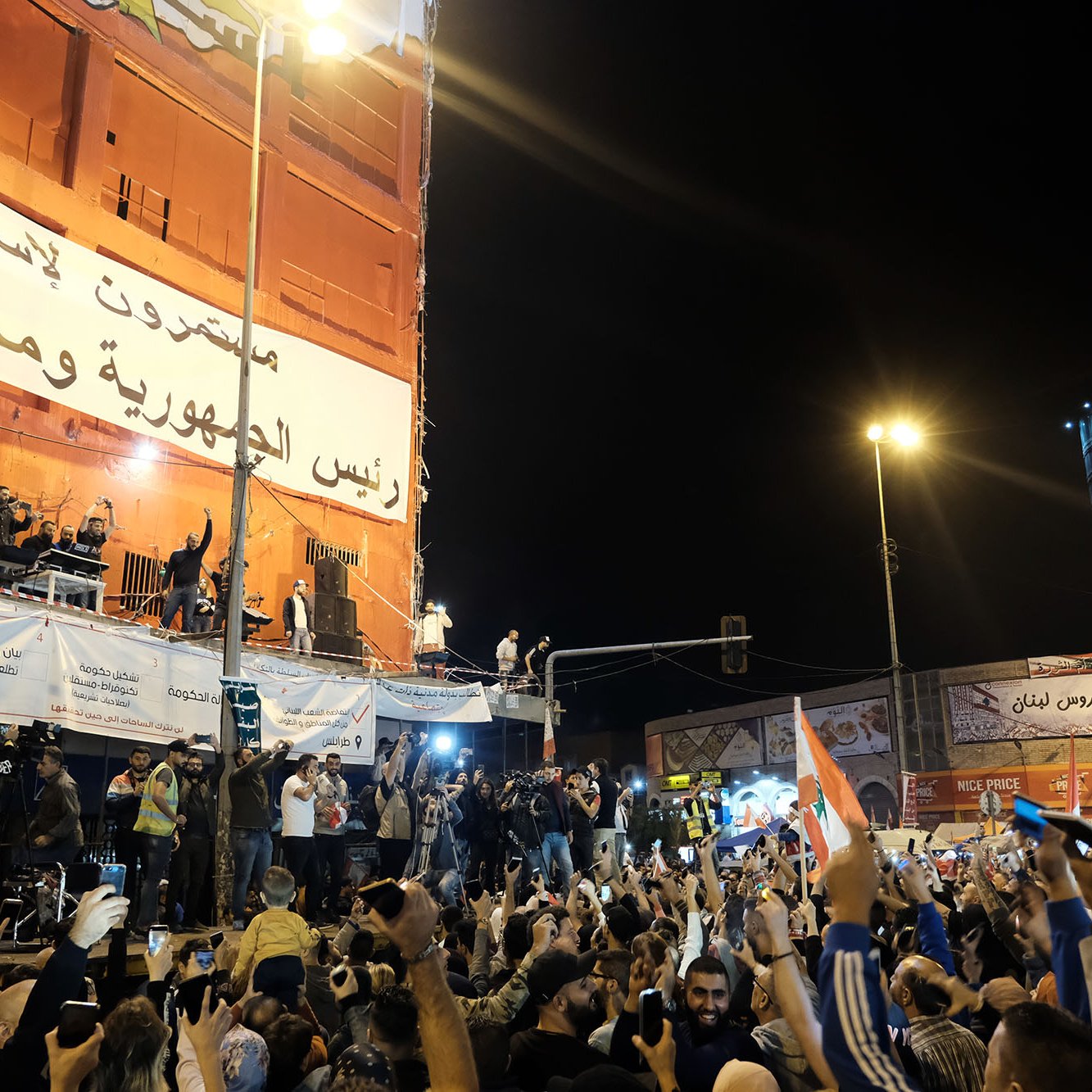 L'image montre une grande foule rassemblée dans une place publique, manifestant avec enthousiasme. Les gens lèvent les mains et brandissent des drapeaux, exprimant des émotions fortes. À l'arrière-plan, on peut voir un grand panneau affichant des messages politiques. Des personnages sont visibles sur une scène surélevée, semblant s'adresser à la foule. L'ambiance semble festive et dynamique, avec des lumières qui illuminent la scène.