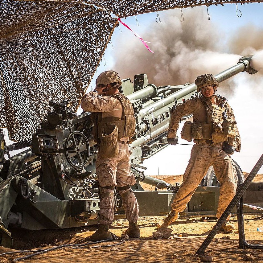 در این تصویر، دو سرباز در یک محیط نظامی دیده می‌شوند که به یک توپخانه بزرگ نزدیک هستند. یکی از سربازان در حال کار با ابزارهای مربوط به توپخانه است، در حالی که دیگری در حال نظارت بر عملیات به نظر می‌رسد. دود ناشی از شلیک گلوله به سمت آسمان می‌رود و پرده‌ای از توری برای حفاظت سربازان قرار داده شده است. فضای اطراف خشک و بیابانی به نظر می‌رسد، که نشان‌دهنده یک عملیات نظامی در یک منطقه جنگی است.