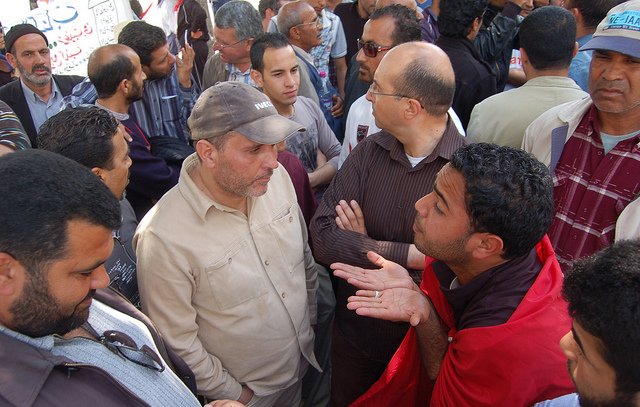 L'image montre un groupe de personnes rassemblées dans un endroit public, probablement lors d'une manifestation ou d'un rassemblement. Plusieurs hommes se tiennent ou se parlent les uns aux autres, affichant des expressions variées qui semblent indiquer une discussion animée. Certains portent des vêtements décontractés et il y a une ambiance de dialogue, avec des gestuelles qui suggèrent l'échange d'opinions. En arrière-plan, on peut apercevoir d'autres personnes qui regardent la scène. L'atmosphère semble engageante et dynamique.
