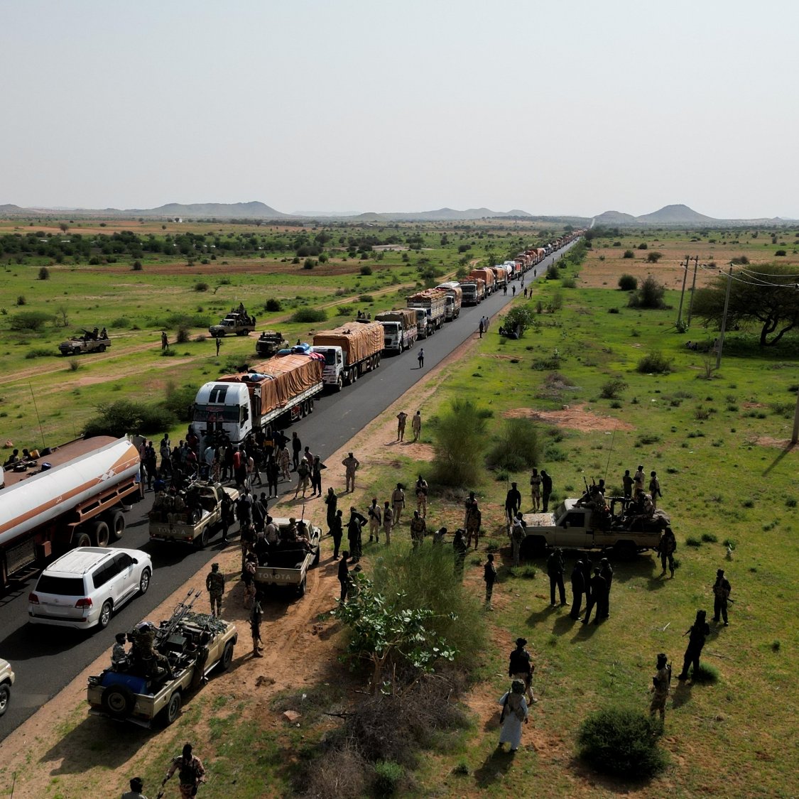 A long line of trucks on a road, with soldiers and green fields surrounding them.