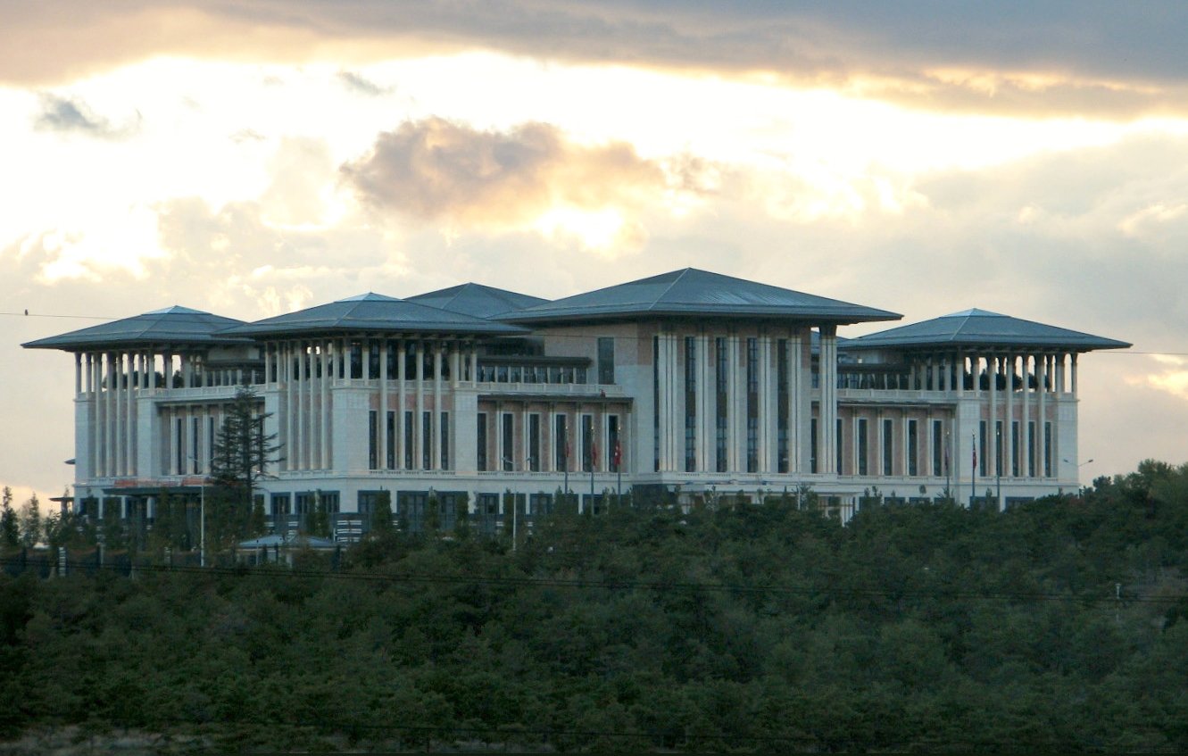 L'image montre un bâtiment impressionnant, probablement un complexe gouvernemental ou administratif. Il possède une architecture moderne, avec plusieurs niveaux et des toits en pente. Les murs sont principalement en blanc, avec de grandes fenêtres, ce qui laisse penser à un design spacieux et lumineux. Le bâtiment est entouré d'un paysage verdoyant, ce qui ajoute à son aspect majestueux. Le ciel en arrière-plan est nuageux, ce qui crée une ambiance dramatique.