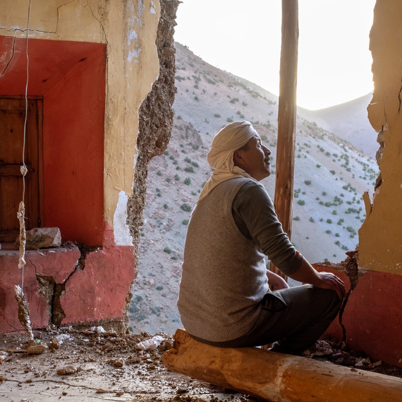 L'image montre un homme assis dans une pièce aux murs endommagés, avec des fissures visibles. À sa gauche, on aperçoit une fenêtre en bois, et à droite, le mur est abîmé, laissant entrevoir l'extérieur. La lumière naturelle pénètre dans la pièce, créant une ambiance mélancolique. L'homme semble contemplatif, perdu dans ses pensées, dans un décor qui suggère l'oubli et la dégradation.
