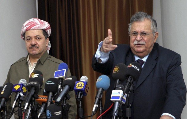 L'image montre deux hommes en conférence de presse, entourés de plusieurs micros et caméras provenant de divers médias. L'homme à gauche porte une tenue traditionnelle kurde avec un foulard, tandis que l'homme à droite, habillé en costume, semble s'adresser à l'audience en pointant du doigt. L'ambiance est formelle, indiquant un moment important de communication ou d'annonce.