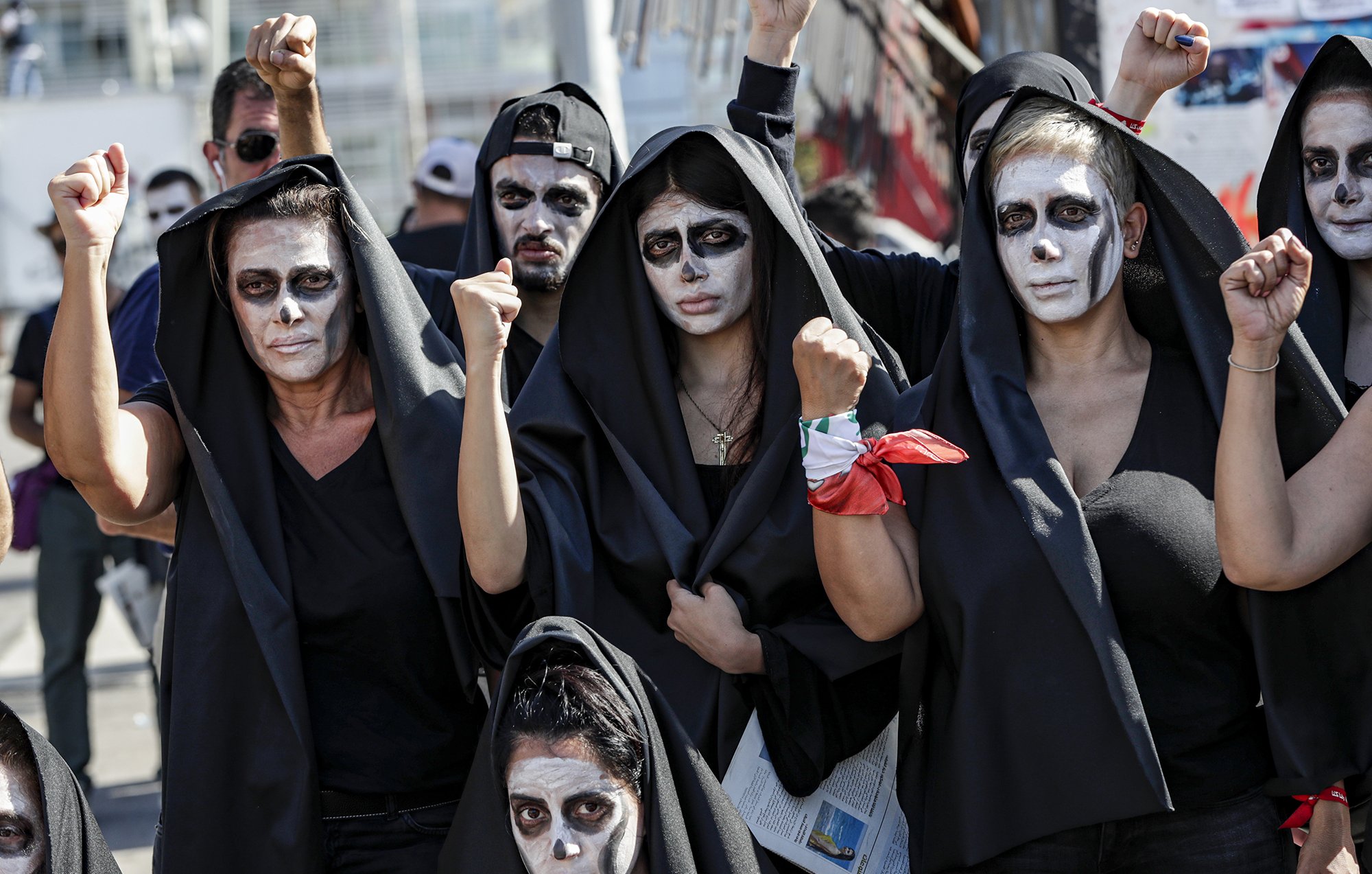 The image depicts a group of people wearing black hooded cloaks and painted faces, featuring white and dark makeup that gives a striking visual effect. They appear to be raising their fists in a gesture of solidarity or protest. The atmosphere suggests they are part of a demonstration or performance, possibly conveying a message or advocating for a specific cause. The backdrop includes hints of a larger gathering, emphasizing the collective nature of their presence.